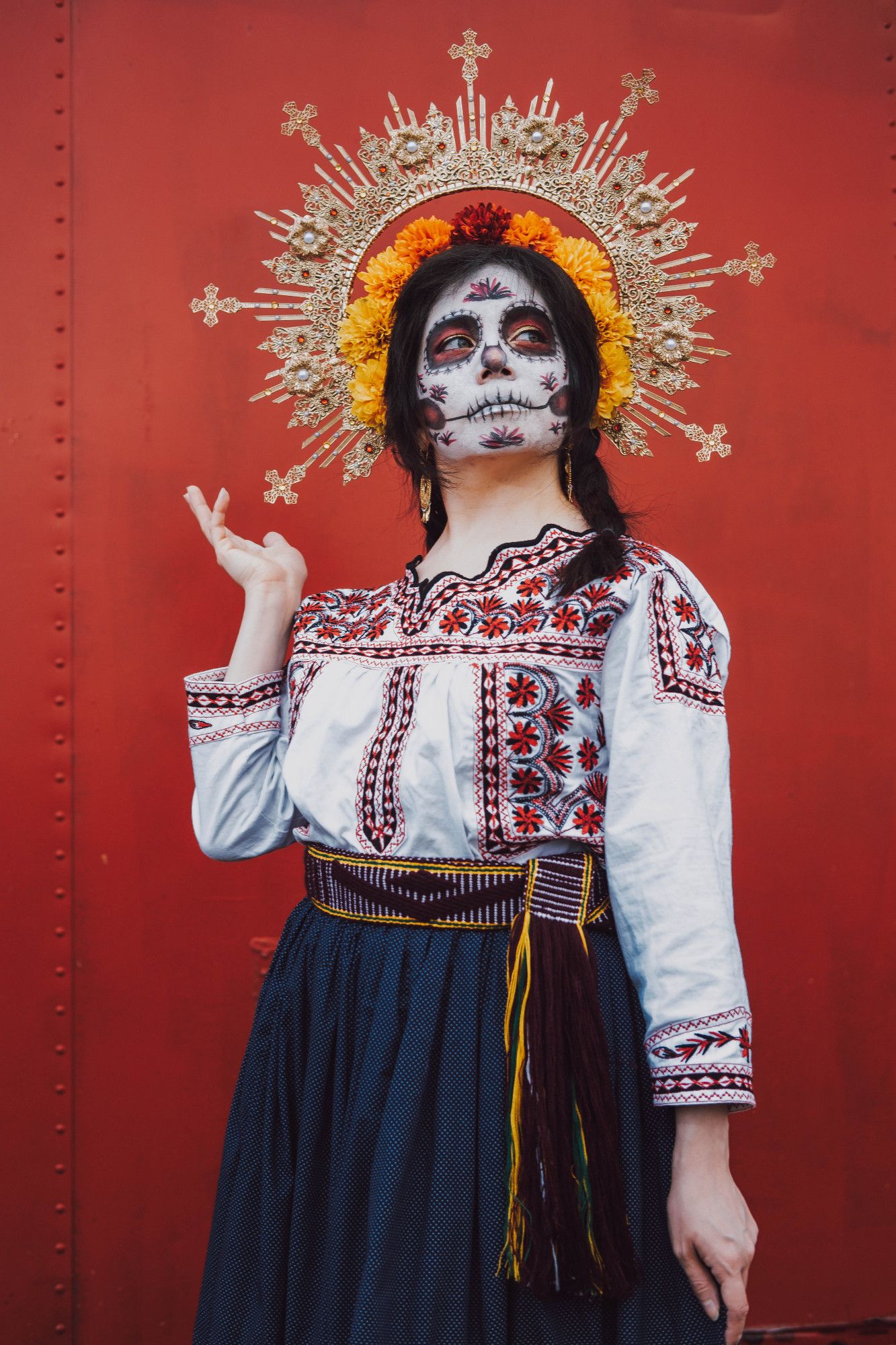 Sophie is standing in front of a solid red background and is looking up. She is wearing a gold halo filigree headpiece with pearls and red rhinestones. There is a halo of red and orange marigolds above her dark hair, which is in two braids. She is wearing gold filigree earrings, a traditional Mexican shirt with red and black flower Mixtec embroidery, a purple and yellow fabric belt, and a blue skirt. Her face is in a Catrina (decorative skull makeup) that has patterns that match the shirt embroidery. One hand is down at her side, and the other is gently framing the edge of her crown.