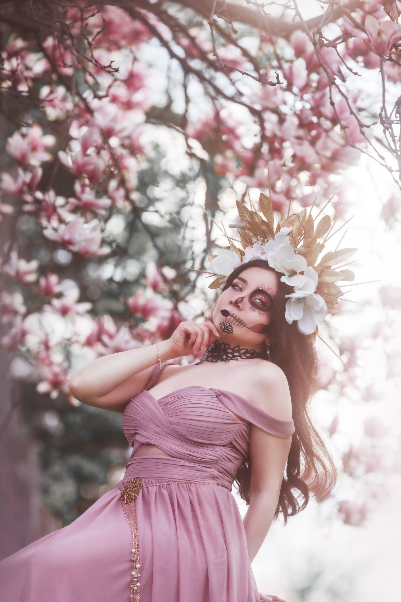 Sophie sitting in front of a blossoming magnolia tree in a graveyard. She is leaning back and supporting herself with one arm; her other hand is at her chin as she appraises the viewer. She is wearing a headpiece of golden leaves, gold spikes, and magnolia flowers. The half of her face that is visible is in pink, gold and black Catrina makeup (decorative skull makeup). She is wearing a large black leather choker in the shape of leaves, a dusty pink dress with off-the-shoulder straps, a thin gold bracelet with pearls, and a delicate gold chain and filigree belt.