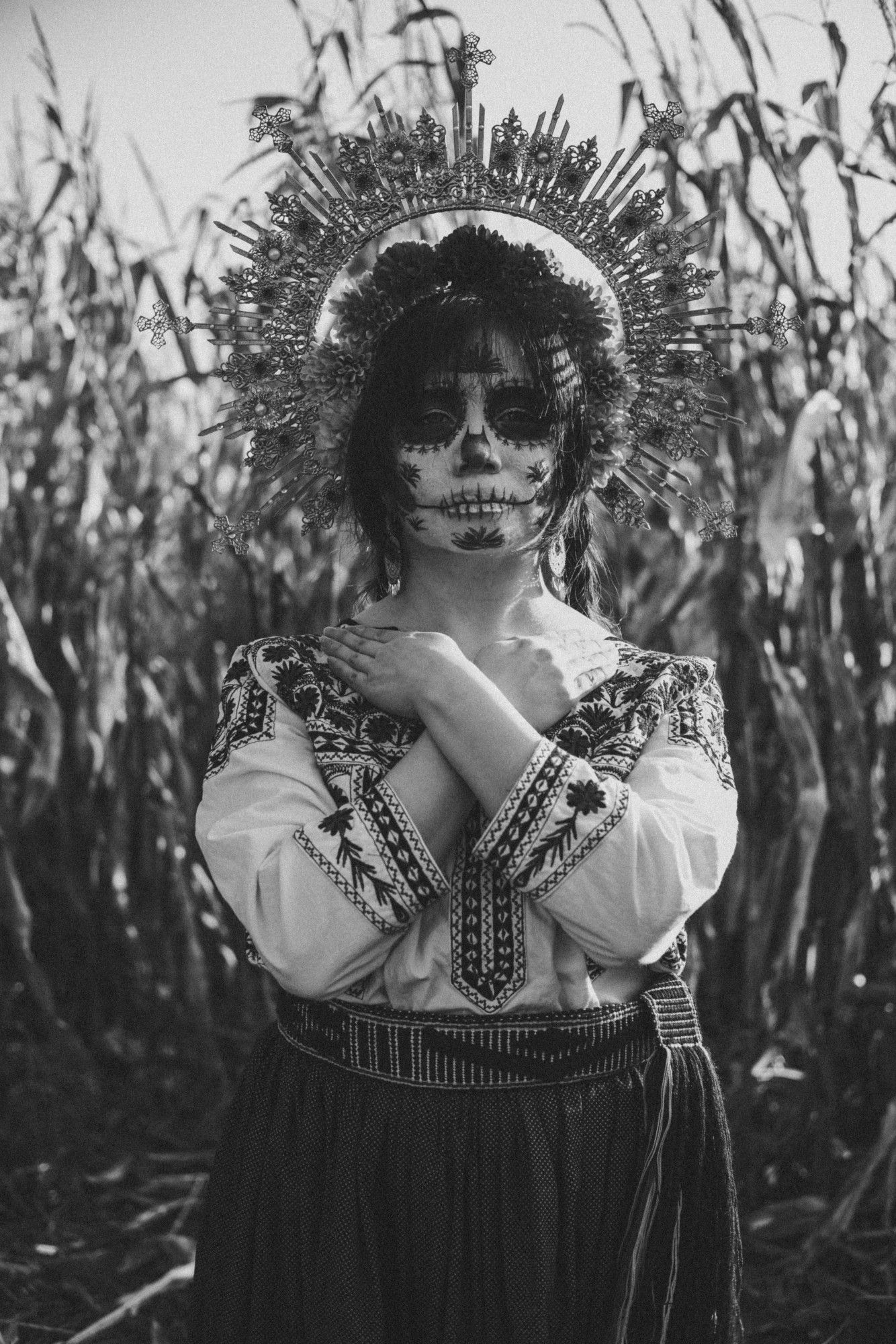 Sophie is standing in a cornfield, smirking at the camera. She is wearing a halo crown covered in filigree and marigolds, a shirt covered in Mixtec embroidery, a fabric skirt, and a fabric belt. Her arms are crossed on her chest with her hands near her neck. Her face is made up in a Catrina (decorative skull makeup) that has designs based on the Mixtec embroidery on her shirt. Her hair is in two braids and she is wearing dangling filigree earrings. The whole photo is in black and white, and there is a bright white flash of light along the right side of the frame.