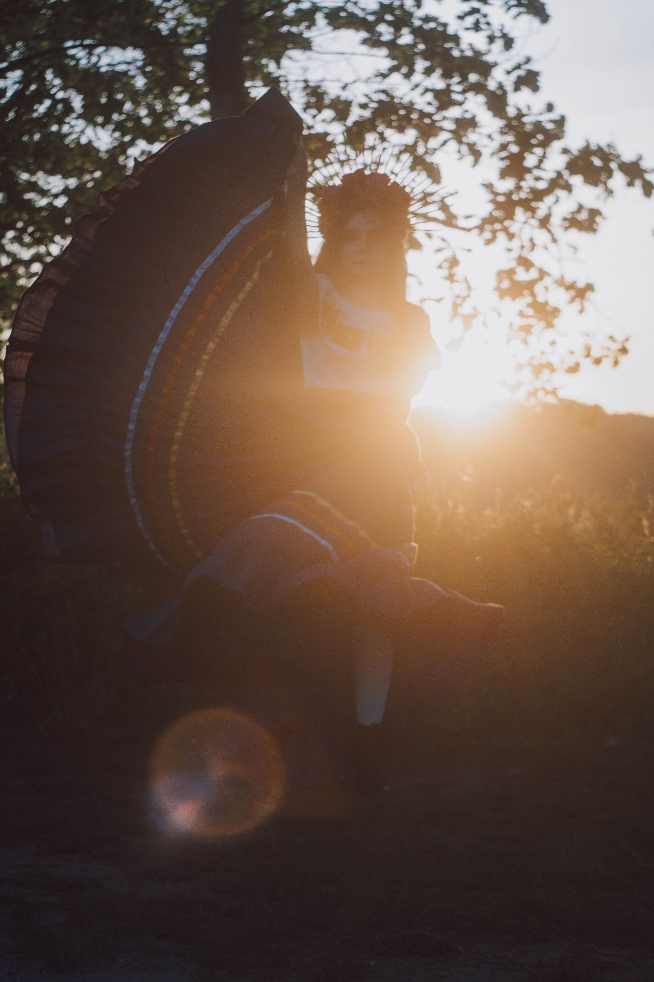 Sophie is standing in shadow with the setting sun shining out at her side. The side of her purple Mexican folkloric dance skirt is arching u at her side. She is wearing a flower headpiece with rays.