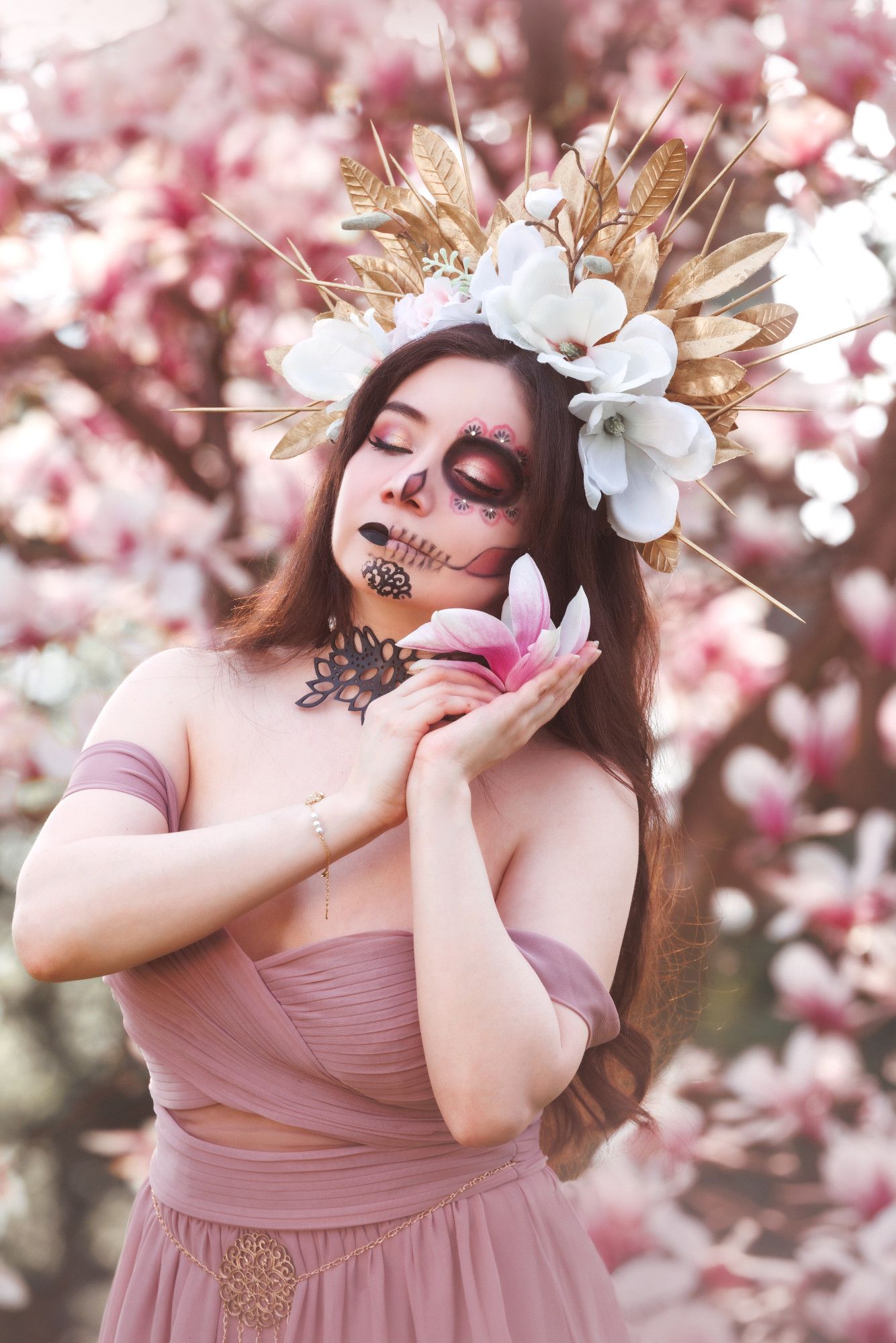 Sophie is closing her eyes and holding a magnolia serenely to her ear. She is standing in front of a blossoming magnolia tree full of pink and white flowers. She is wearing a headpiece of golden leaves, gold spikes, and magnolia flowers. Half of her face is in normal makeup with pink and gold eyeshadow and black lipstick. The other half of her face is painted in a Catrina (decorative skull makeup) using the same makeup colors. She is wearing a large black leather choker in the shape of leaves, a dusty pink dress with off-the-shoulder straps, and a gold chain and filigree belt.