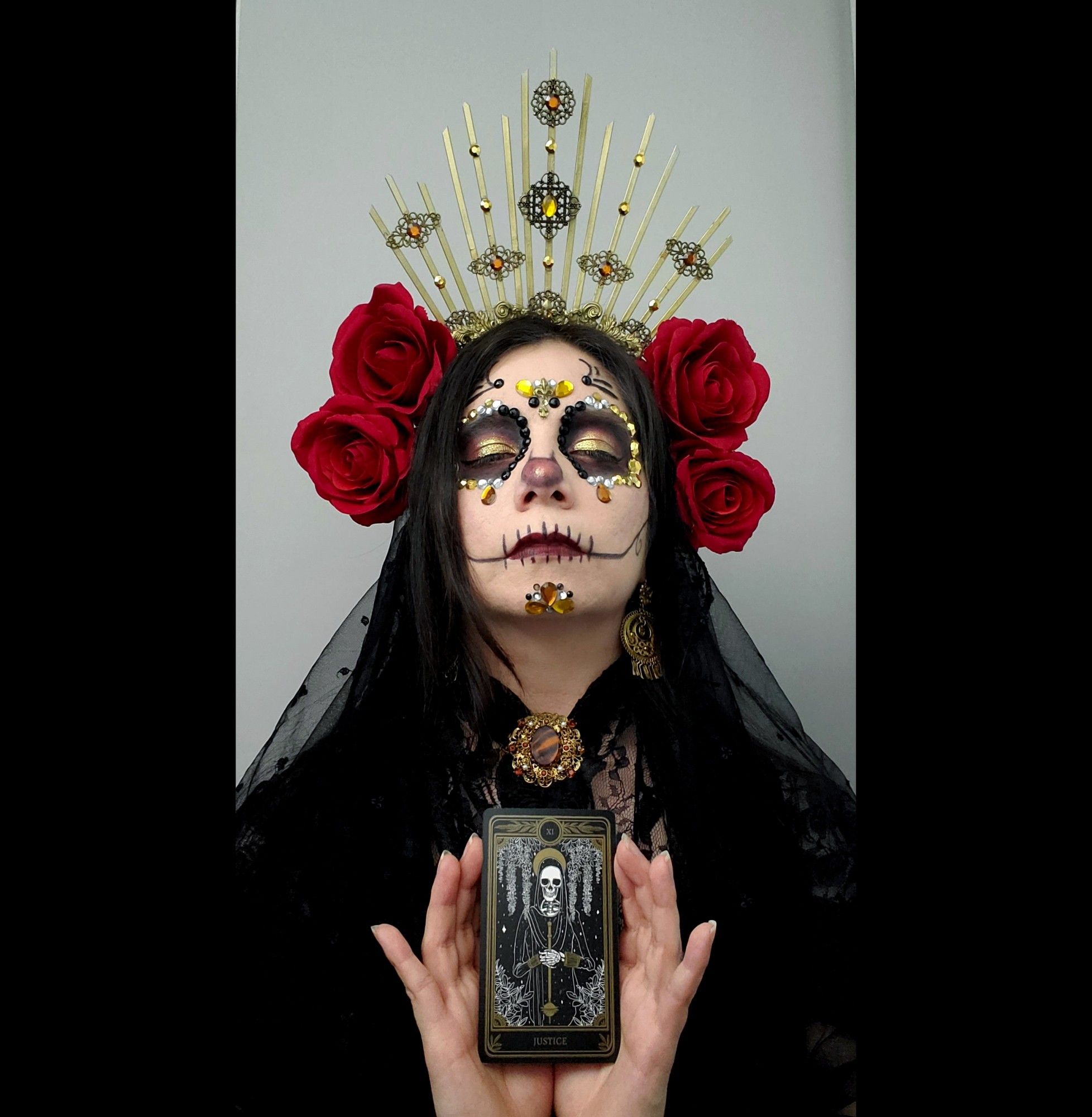 Sophie is in front of a black background wearing a black lace top and a black translucent veil over her dark hair. She is wearing a gold ray headpiece with filigree and yellow and orange jewels. There are two large red roses on either side of her face. She is wearing a Catrina (decorative skull makeup) made up of black, red, and gold makeup, as well as many black, yellow, orange, and white jewels. She has on gold filigree earrings and a dark orange filigree brooch at her neck. Her eyes are closed and she is holding a skeleton Justice tarot card, which is done in a black and white Punjabi style with gold trim.