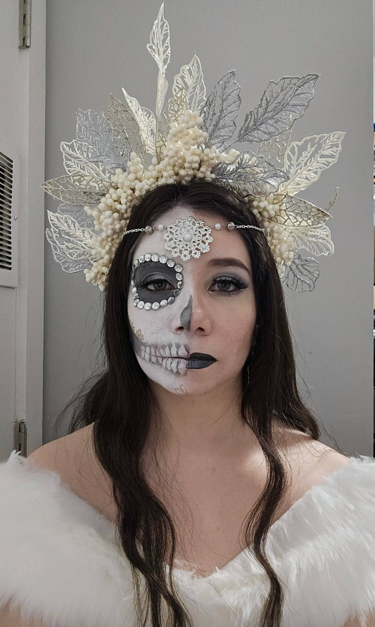 Sophie is staring into the camera with an uncaring expression. She has pale skin and long, dark, wavy brown hair. She is wearing an off-the-shoulder white fur top and a headpiece made of glittering silver, white, and gold leaves and cream-colored berries. There is a silver chain with silver beads and pearls holding up a large, circular piece of filigree over her forehead. Half of her makeup is done in glamorous silver and grey eyeshadow, sharp cheekbone contour, and dark grey metallic lipstick. The other half of her face is in a white Catrina (decorative skull makeup) with the hollows of her eyes, nose, and cheekbone is silver and grey. Her eye socket is lined in silvery-clear rhinestones, and there are teeth painted over her lips. Small, gold flowers are drawn on her chin and along her cheekbone.