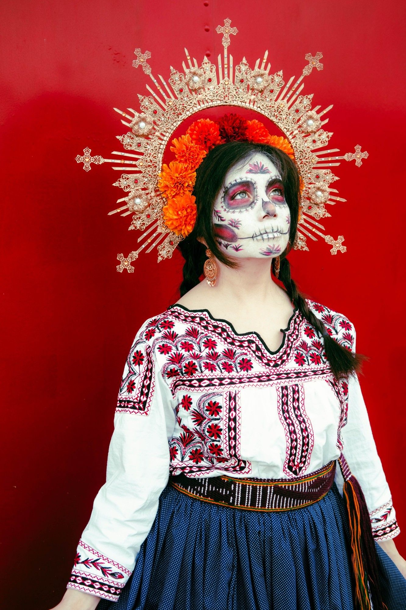 Sophie is standing in front of a solid red background and is looking up. She is wearing a gold halo filigree headpiece with pearls and red rhinestones. There is a halo of red and orange marigolds above her dark hair, which is in two braids. She is wearing gold filigree earrings, a traditional Mexican shirt with red and black flower Mixtec embroidery, a purple and yellow fabric belt, and a blue skirt. Her face is in a Catrina (decorative skull makeup) that has patterns that match the shirt embroidery.