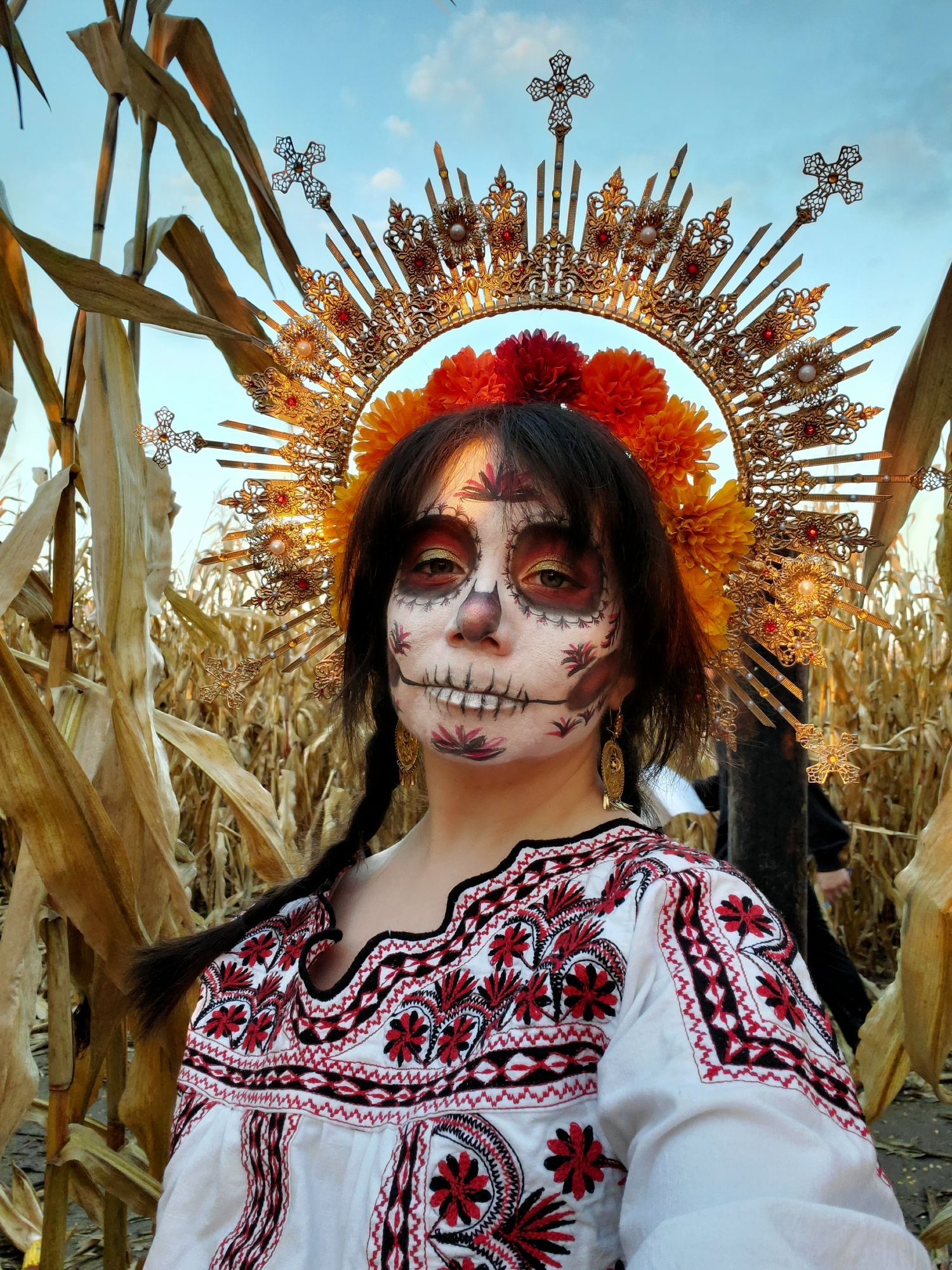 Sophie is outside in the middle of a corn maze with the sun's rays dappled over her. She has on a large gold halo headpiece with filigree, red rhinestones, and pearls. There is a smaller halo of red, orange, and yellow marigolds close to her hair, which is dark brown and in two long braids. She is wearing gold filigree earrings and a white traditional Mexican shirt in black and red Mixtec embroidery. She has pale skin and her face is painted in a white Catrina (decorative skull makeup) with red, gold, and black decorations that match the embroidery.