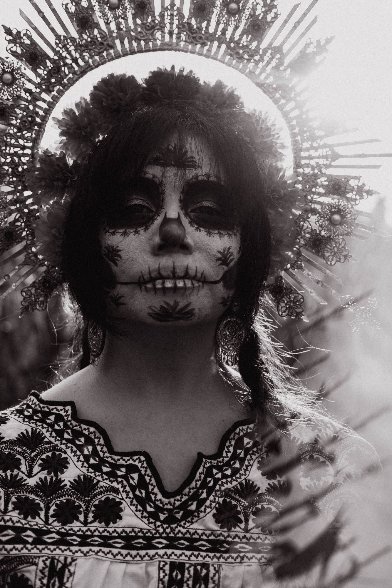 Sophie is standing in a cornfield, smirking at the camera. She is wearing a halo crown covered in filigree and marigolds and a shirt covered in Mixtec embroidery. Her face is made up in a Catrina (decorative skull makeup) that has designs based on the Mixtec embroidery on her shirt. Her hair is in two braids and she is wearing dangling filigree earrings. The whole photo is in black and white, and there is a bright white flash of light along the right side of the frame.