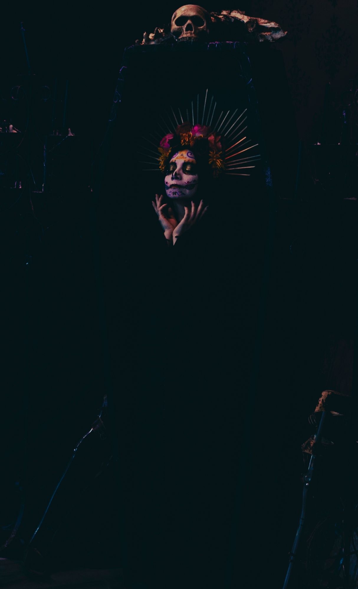 Sophie is wearing black and fading into the black coffin she is standing in. The coffin has a skull and bones on top. Her hands are clawed gently around her face. Her eyes are closed and she is in colorful Catrina makeup (decorative skull makeup). She is wearing a bright rose and marigold flower crown with gold rays.