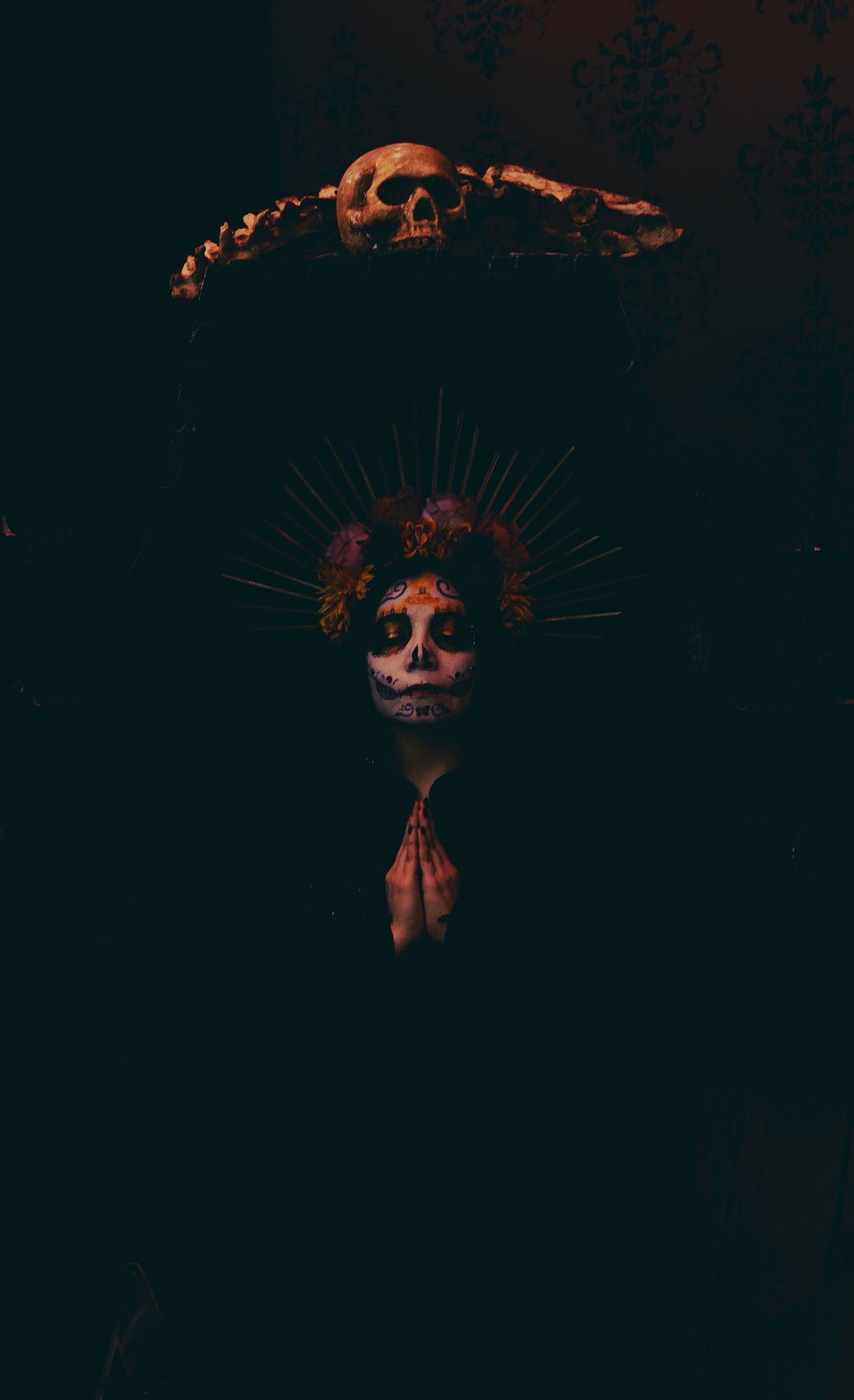 Sophie is wearing black and fading into the black coffin she is standing in. The coffin has a skull and bones on top. Her pale hands are pressed in prayer at her chest. Her eyes are closed and she is in colorful Catrina makeup (decorative skull makeup). She is wearing a bright rose and marigold flower crown with gold rays.