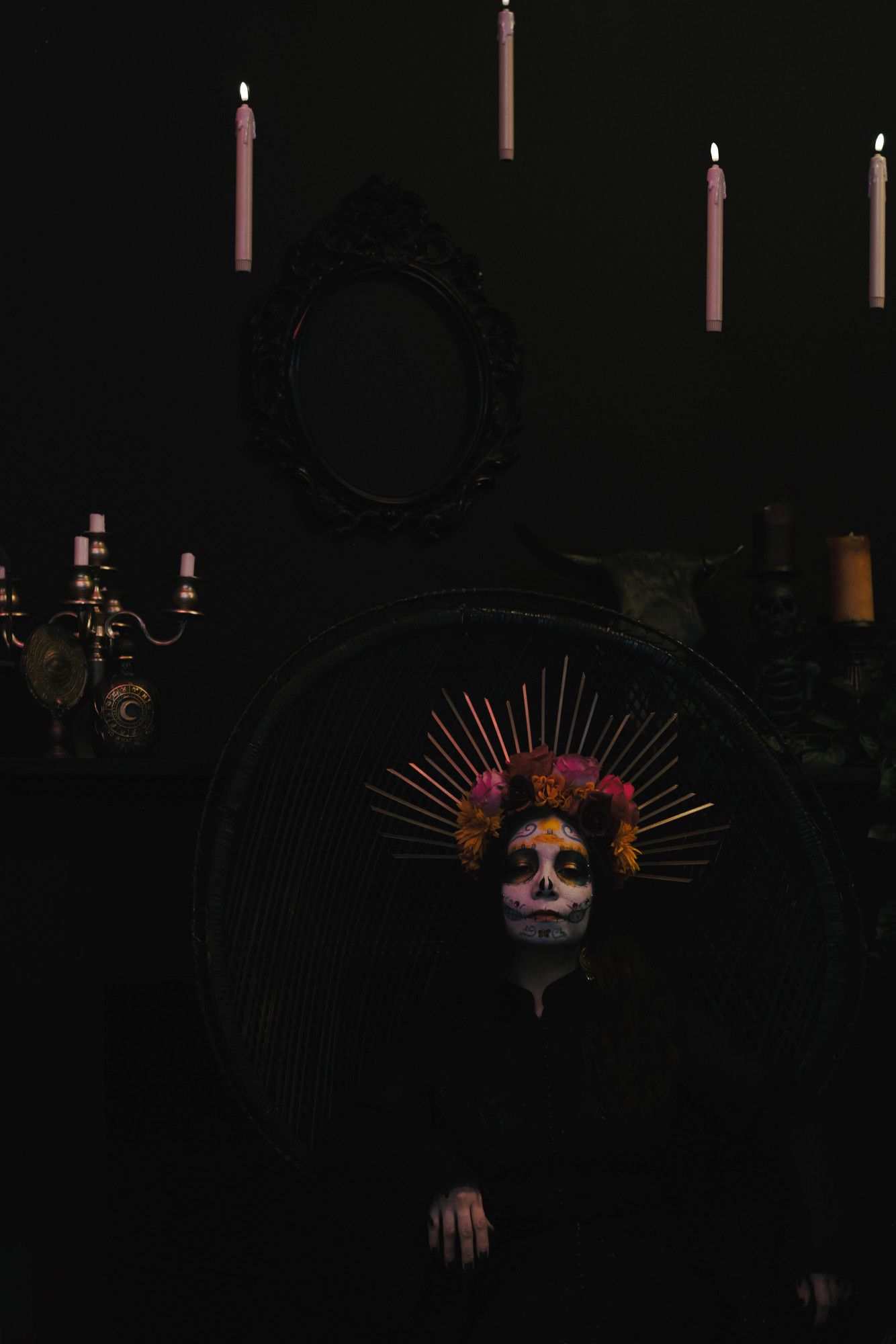 Sophie is wearing black and is fading into the black wicker chair she is sitting in. She is in front of a black fireplace and matching black wall. There is a golden candelabra, a cow skull, and a black empty picture frame behind her. Candles float above her head. Her eyes are almost closed and she is in colorful Catrina makeup (decorative skull makeup). She is wearing a bright rose and marigold flower crown with gold rays. Her pale hands are seen resting on the chair arms, and her dark hair fades into the background. She looks vaguely uninterested in the viewer.