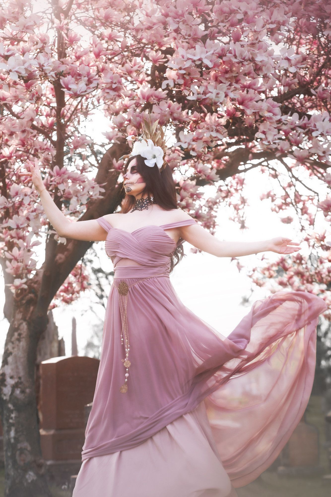 Sophie is standing in front of a blossoming magnolia tree in a graveyard. She is looking to the side with outstretched arms; her skirt is flying behind her in the wind. She is wearing a headpiece of golden leaves, gold spikes, and magnolia flowers. The half of her face that is visible is in pink, gold and black Catrina makeup (decorative skull makeup). She is wearing a large black leather choker in the shape of leaves, a dusty pink dress with off-the-shoulder straps, and a delicate gold chain and filigree belt.