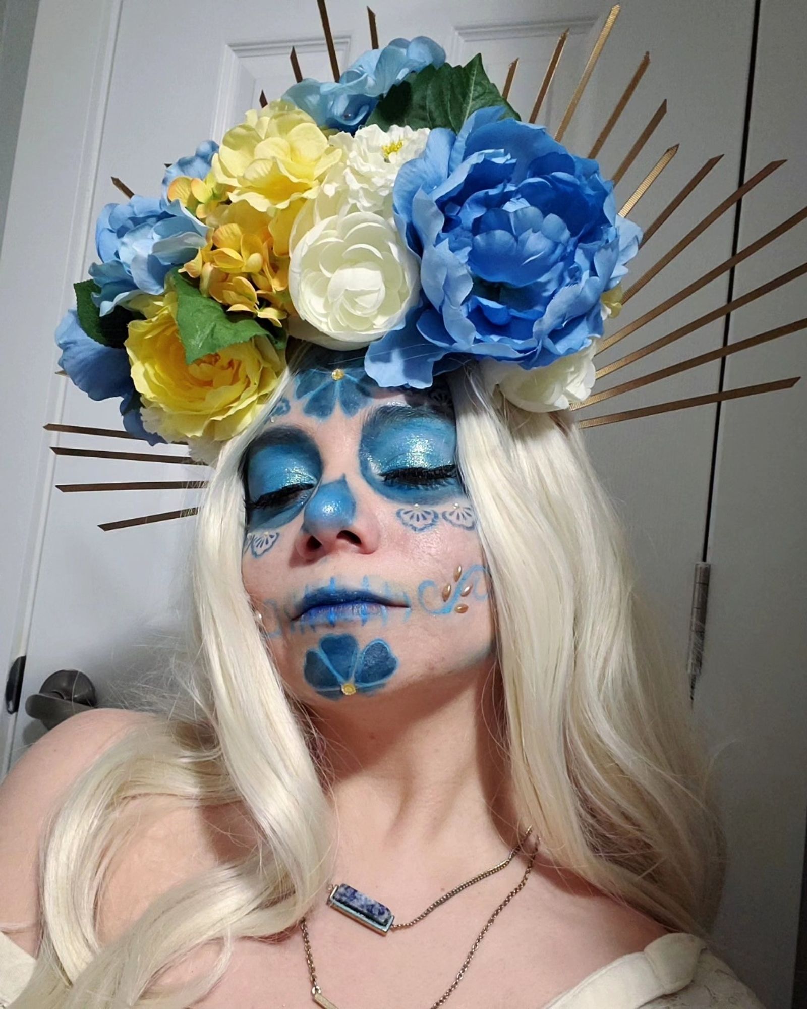 Sophie is wearing a colorful blue, white and yellow flower headpiece with gold rays. Her face is painted in a monochromatic blue Catrina (decorative skull makeup). She has on a white shirt with a blue necklace. Her skin is pale and her hair is long and a wavy white blonde. The background is a plain white wall.