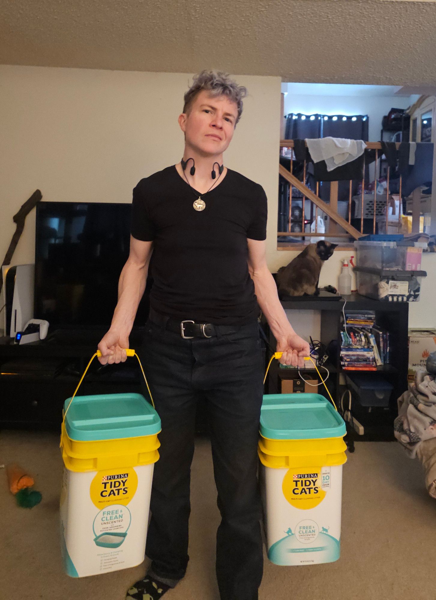 White genderqueer person with messy curly silvery blue hair and stormy blue eyes wearing a black tshirt and dark blue jeans with a leather belt, holding a 35lb cat litter bucket in each hand and casually flexing arms with powerful vein structure while they look over at the camera with an intense almost smirk