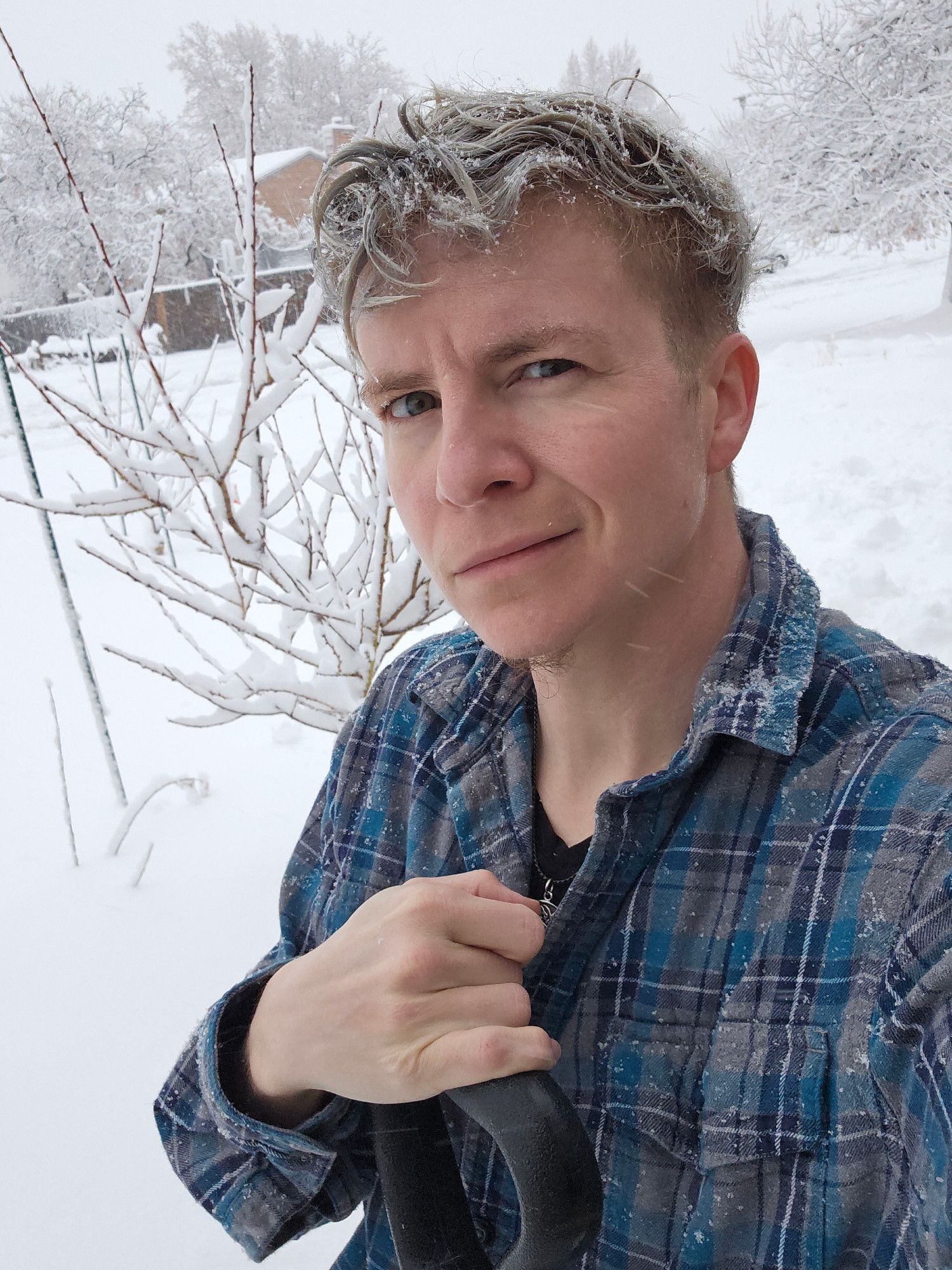 White genderqueer person with messy curly silvery blue hair and stormy blue eyes, wearing a blue and grey flannel and leaning on a snow shovel, smirking in gently commanding flirtation while snow catches in their hair