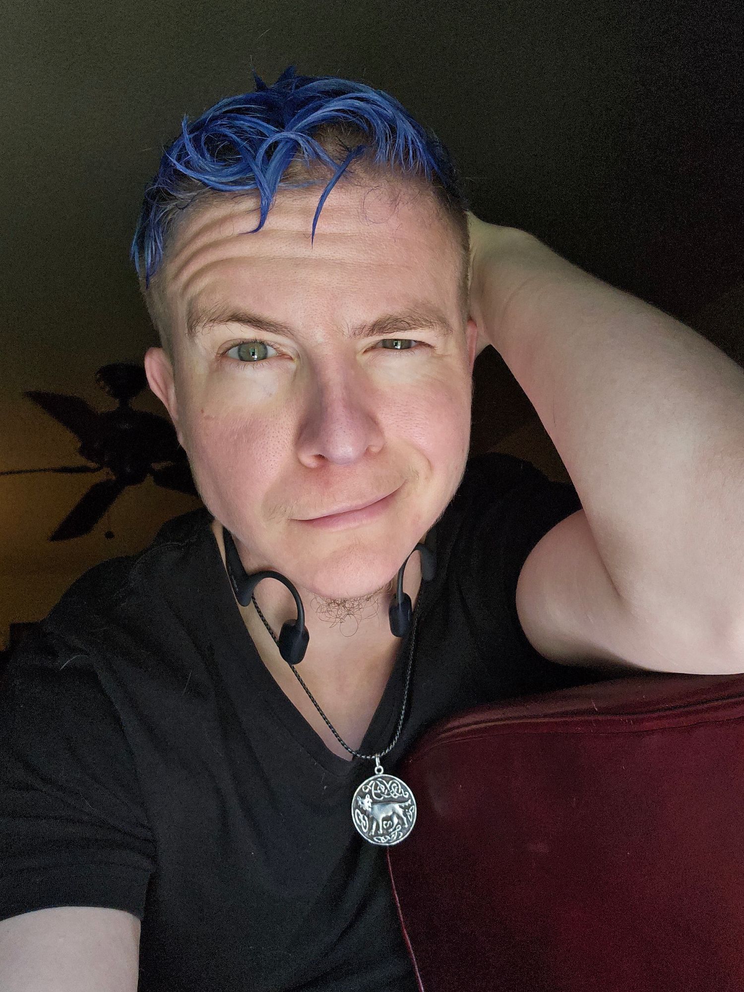 White genderqueer person with messy curly cobalt blue hair and stormy blue eyes wearing a black tshirt, leaning on one arm and smirking softly over at the viewer