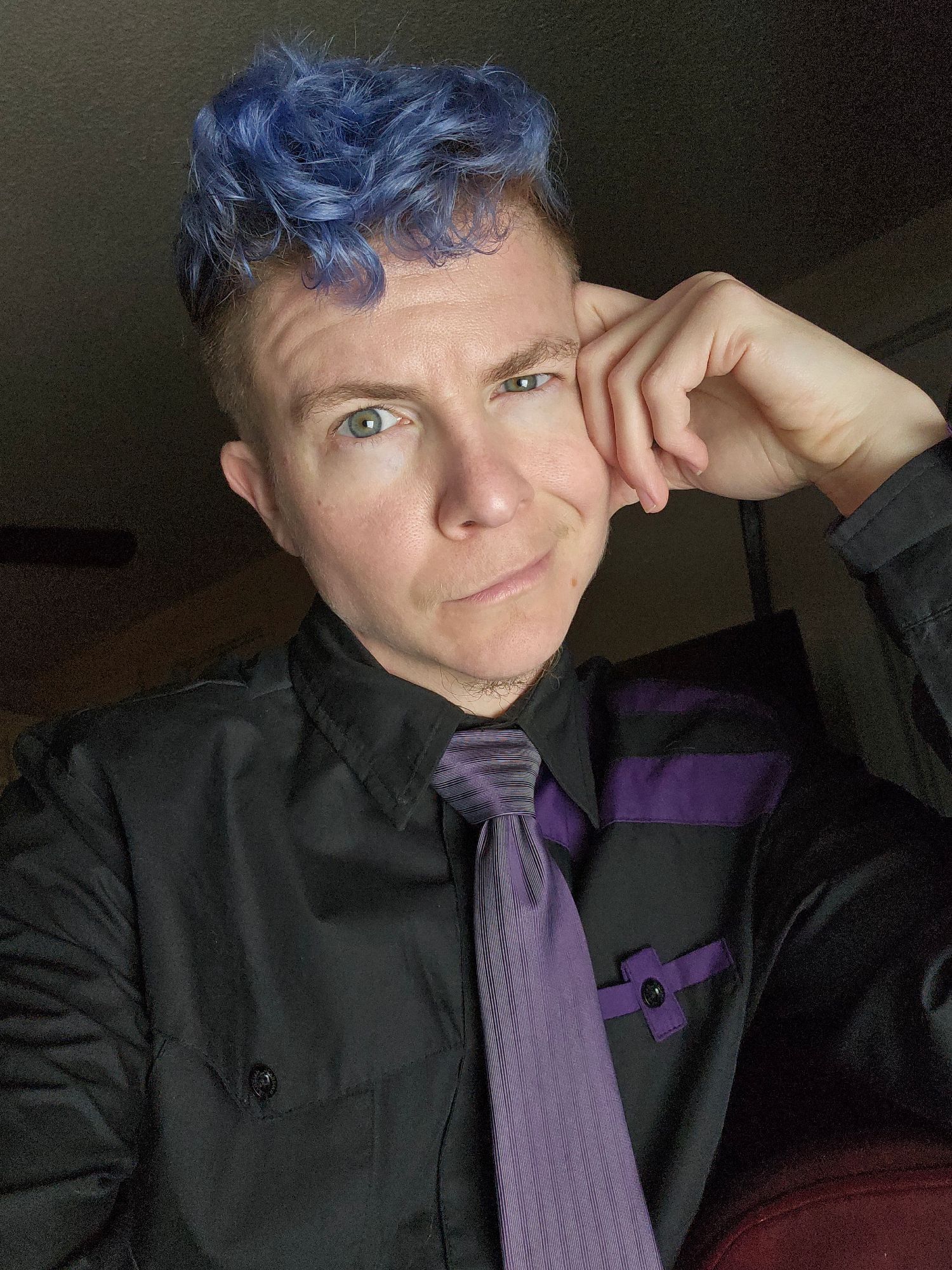 White genderqueer person with messy curly cobalt blue hair and stormy blue eyes wearing a black and purple button up shirt with a purple striped tie, leaning on one hand and smirking in playful invitation