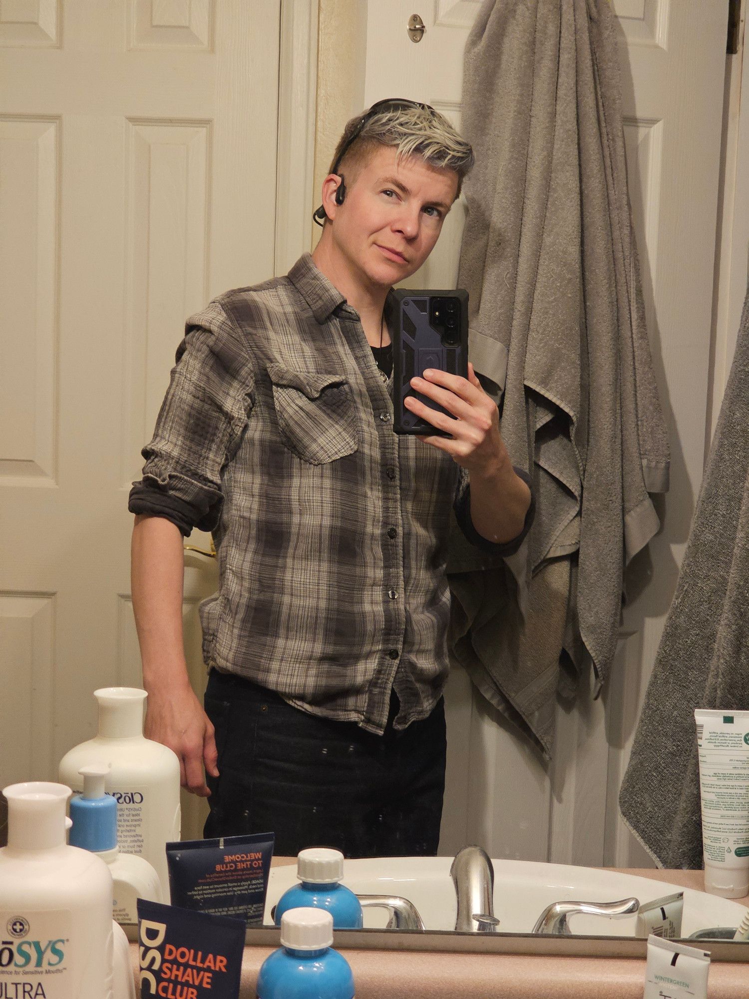 White genderqueer person with messy curly silvery blue hair and stormy blue eyes wearing a grey flannel and dark blue jeans, standing in front of a mirror in which they are taking their picture, smirking in cocky playfulness at the reflection