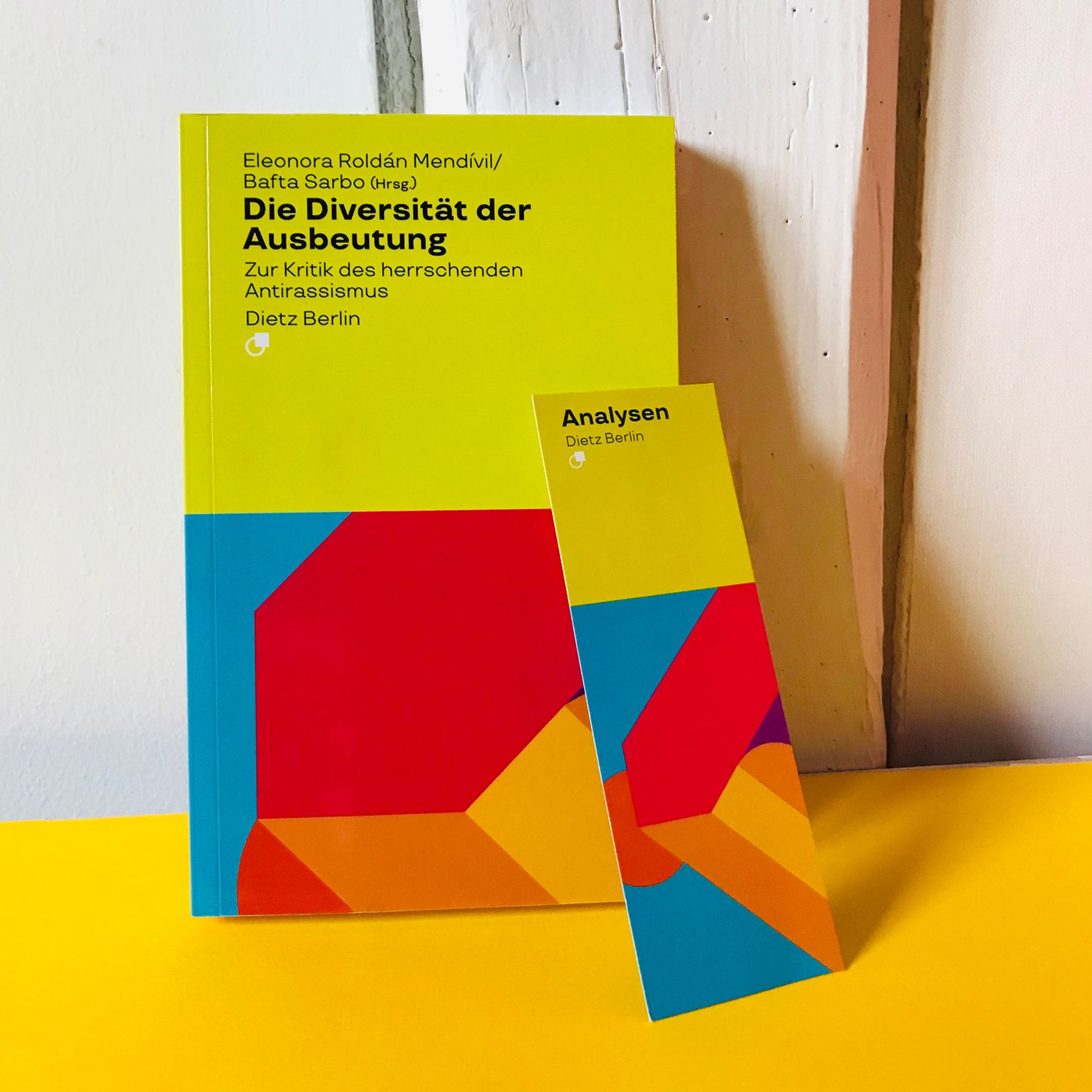 Neben unserem Analysen-Lesezeichen in gleicher Optik steht an die Wand gelehnt der Sammelband „Die Diversität der Ausbeutung. Zur Kritik des herrschenden Antirassismus“, herausgegeben von Eleonora Roldán Mendívil und Bafta Sarbo