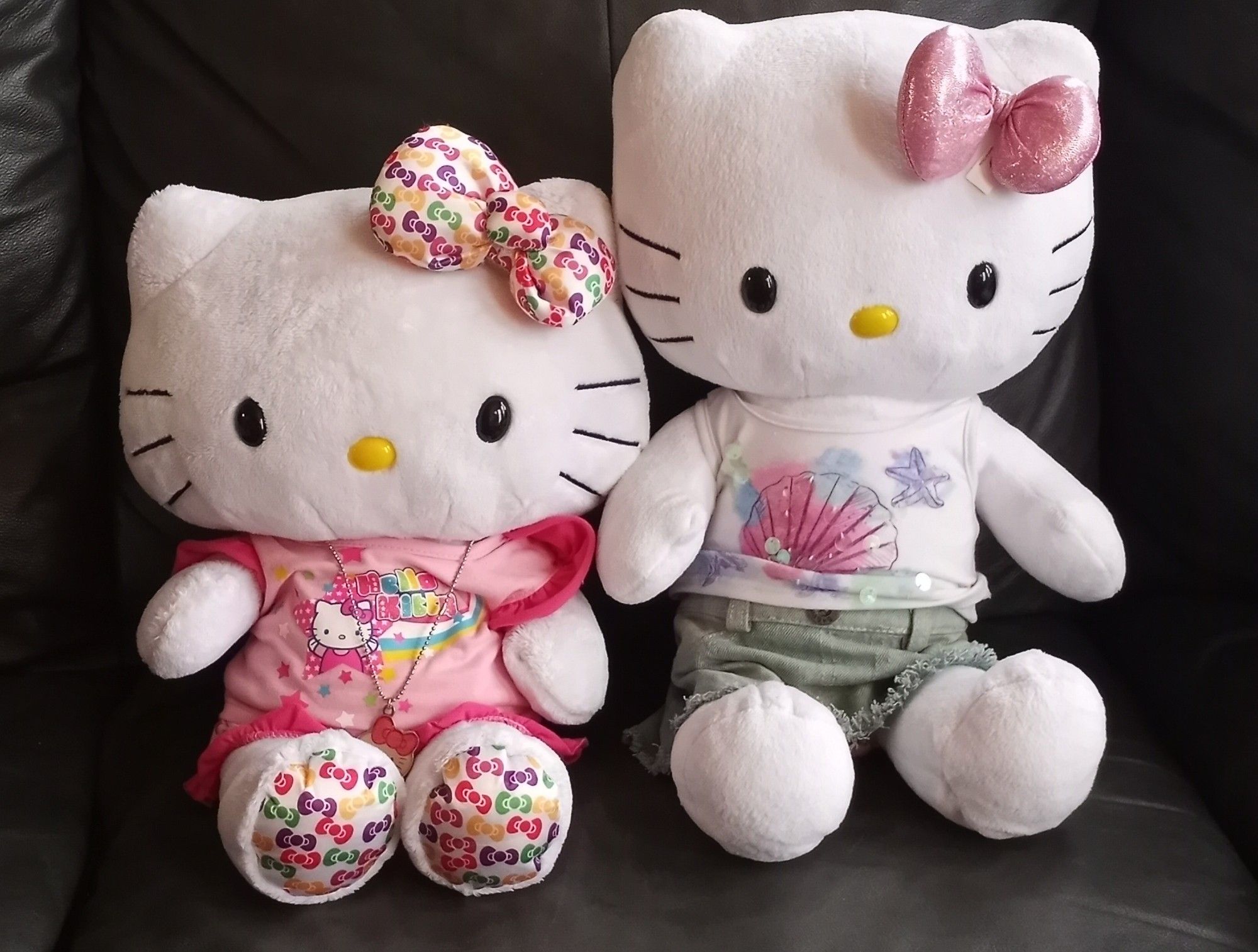 Two Hello Kitty Build-a-Bear plusses. The one on the left for created for HK's 35th Anniversary. Her bow has different colored bows (red, yellow, pink, purple, green) against a white background. The same multi-colored bow print is on the soles of her feet. The one on the right is the standard BABW Hello Kitty, with a light pink shiny bow, a white tank top with a colored shell and starfish, and a light blue denim skirt.