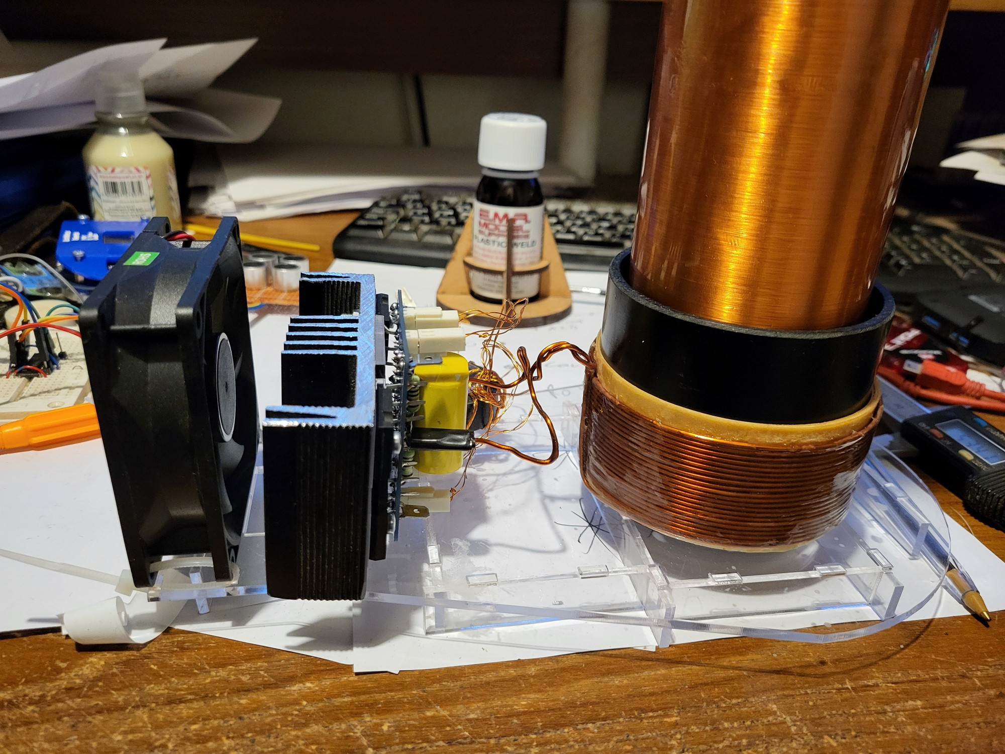 Tesla coil driver left to right. 

Black fan. Large black heat sink with a pcb and 4 transistors 

Primary coil 15 turns of enamelled copper wire and the base of the secondary coil. 