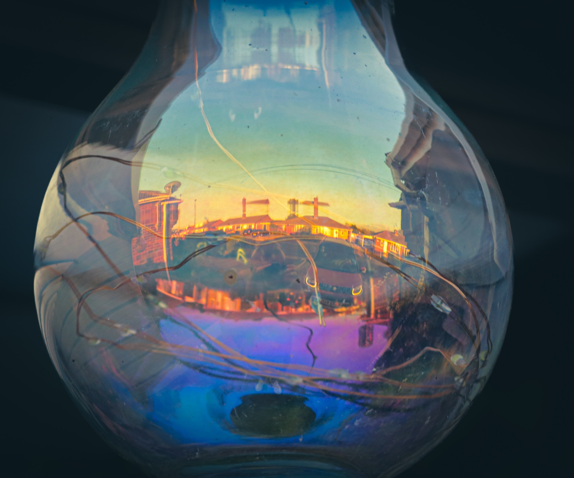 The image depicts a close-up, abstract view of a clear, pear-shaped glass object, likely a light bulb or a decorative glass vessel, with a vibrant reflection within it. The glass surface shows subtle iridescent hues, particularly a prominent blue and purple shimmer at the bottom. Inside the glass, thin, dark, wire-like strands of filaments are visible, seemingly entangled or arranged in a loose pattern.
The most striking feature is the detailed reflection captured within the glass, which appears to be a street scene at either sunrise or sunset. The reflection shows a row of buildings, possibly houses or shops, illuminated by warm, golden light, suggesting a low sun angle. A vehicle, likely a car, is visible in the foreground of the reflection, and there are what appear to be power lines or utility poles against the sky. The sky in the reflection transitions from a bright yellow or orange near the horizon to a lighter blue higher up. The overall background of the image outside the glass object is dark and out of focus, emphasising the central glass element and its internal reflection.