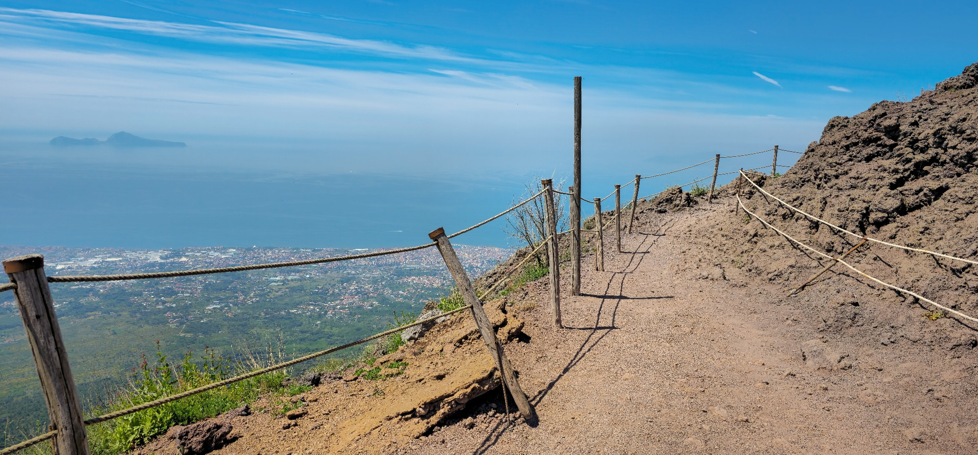 The image depicts a view from the slopes of Mount Vesuvius, looking out over the Bay of Naples and the surrounding landscape.
Foreground:
A dirt path with a rope barrier supported by wooden posts runs along the edge of the volcanic terrain, guiding visitors. The ground is rough and rocky, characteristic of volcanic slopes.
Midground:
Beyond the path, the sprawling urban area of Naples and its surrounding towns are visible, stretching along the coastline. The blue waters of the Bay of Naples extend further out.
Background:
In the distance, two small islands can be seen as a distinct landmass rising from the sea under a clear, bright blue sky with a few wispy clouds. The overall impression is one of a vast, elevated perspective offering a panoramic view of the Campania region.
