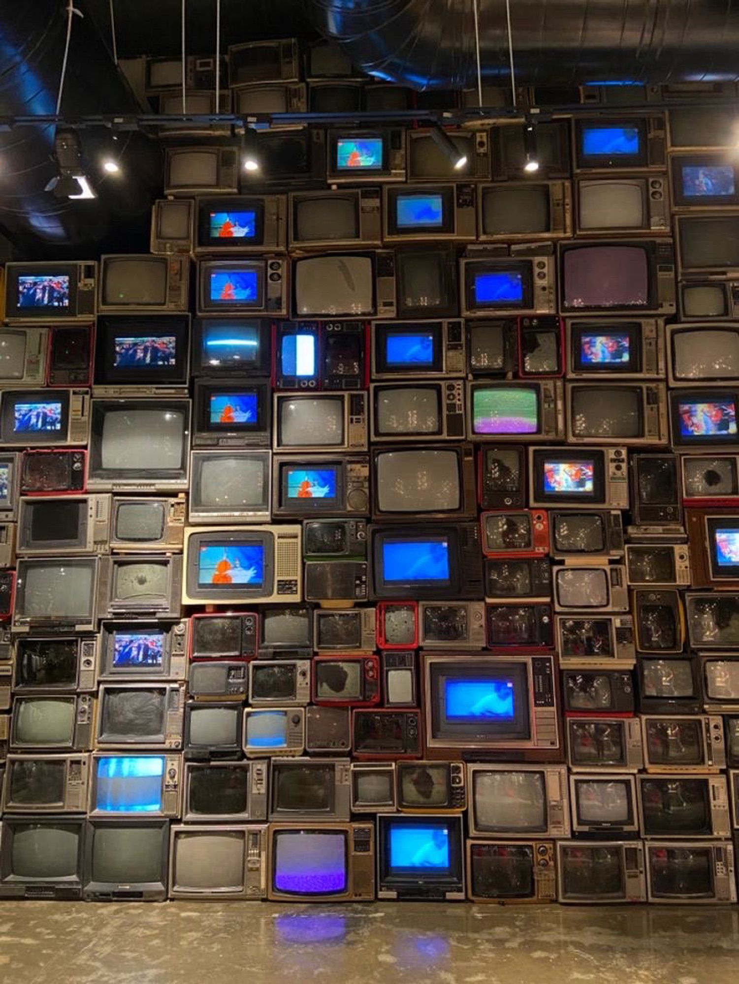 A wall of old TVs stacked on top of each other. Some are turned on and others are not, and some are broken.