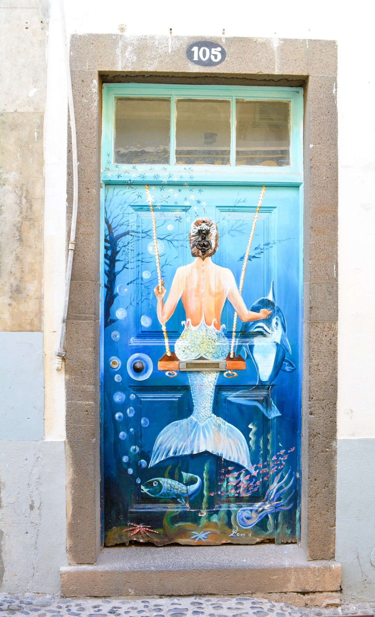 The back of a mermaid on a swing is painted on a door that appears to be under water. She is petting a dolphin. Fish and pupples as well other sea creatures feature near the bottom. There are three glass panels at the top above which is the street number, 105.