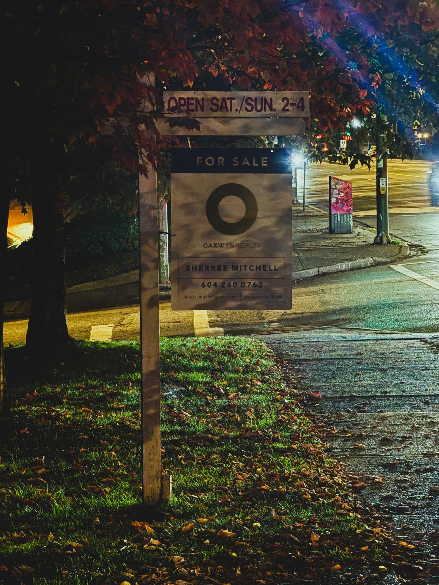 A for sale sign for a condo on a wooden post planted in the grass next to a sidewalk both filled with leaves from fall. A very large O dominates the sign representing the real estate company name - Oakwyn Realty - which is printed in smaller letters underneath. There is a junction just beyond it with a red mailbox and the lights of oncoming traffic.