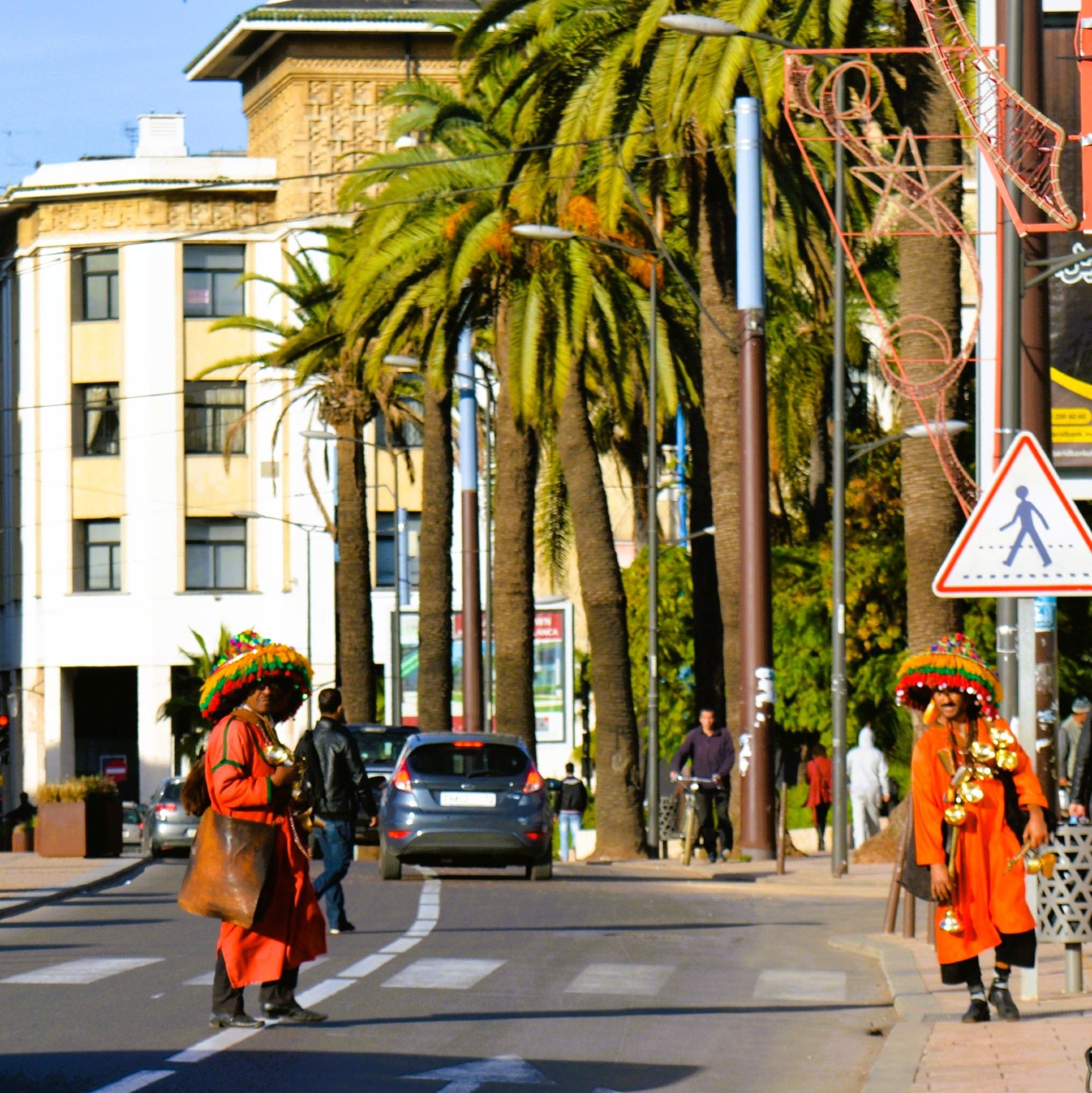 Two garrabs wearing red djellaba, and matching large ornate hats that look like your great aunt Ethel's lampshade standing in the middle of a crosswalk on a Casablanca street. They carry goatskin bags with cold fresh water, and brass cups to serve it. There are pedestrians, cars and cyclists amid palm trees and buildings.