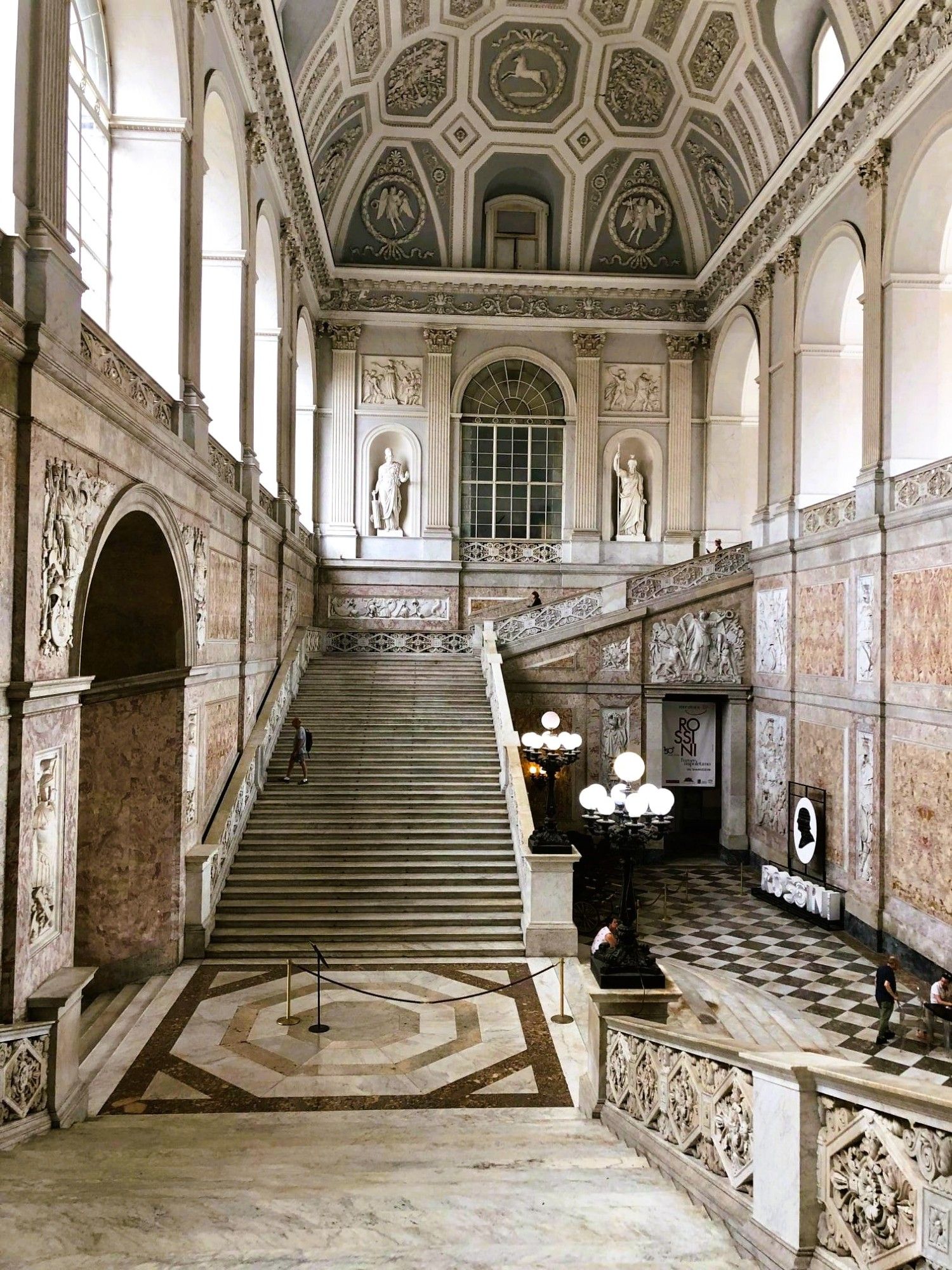 One side of a sweeping marble staircase inside a palace. There are statues in alcoves in the walls, lamp posts with multiple globes, as well ornate decor on the walls and ceiling, and large arched windows letting in the light.