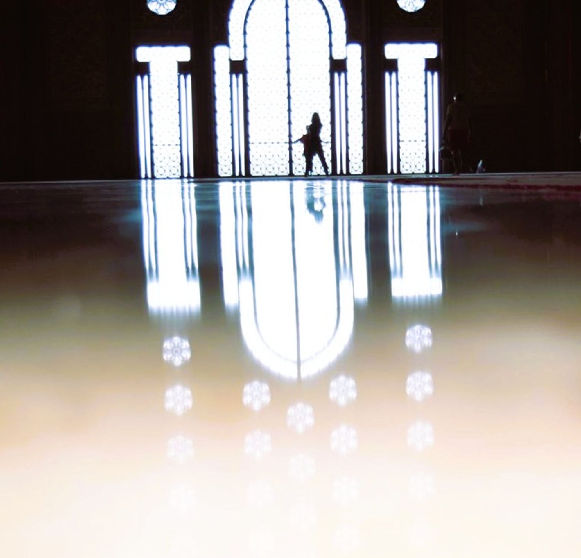 A silhouette of a woman hugging a child as they pose in front of an ornate arched window with two windows on either side that look like pillars. The walls around it are black and so the light is dominant. All windows go down to ground level and their shapes reflect on a highly polished marble floor which also reflects numerous small blossom shaped windows partially seen at the top of the larger windows.