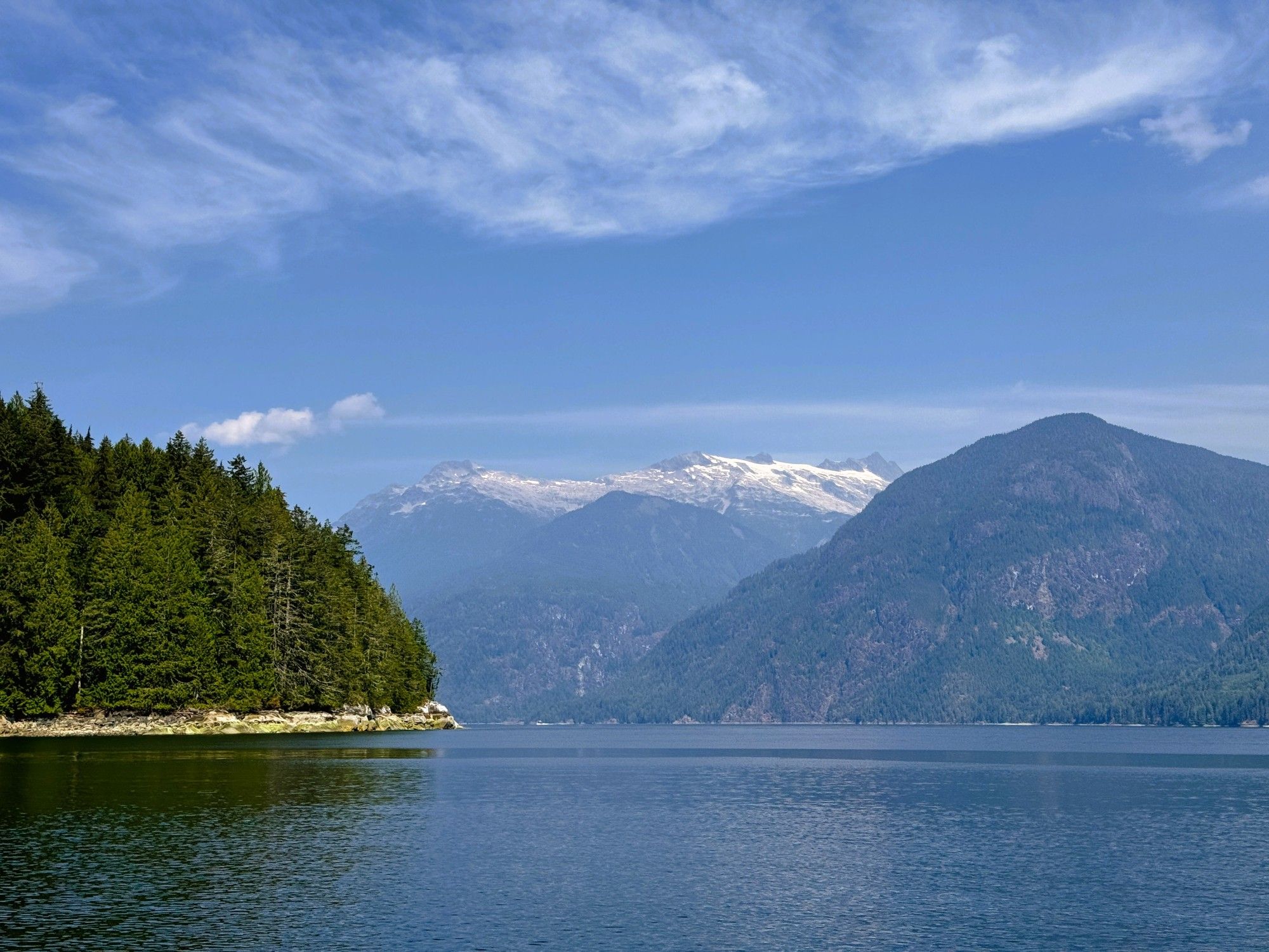 Beautiful vista from the middle of the ocean. On the left is a mountain filled with fir trees and on the right a slightly further out mountain, of rock and trees and in the middle in the far distance a glacier, with snow capped peaks. A blue and slightly white cloudy sky reflects on the calm of the ocean. Overall a Canadiana wilderness scene.