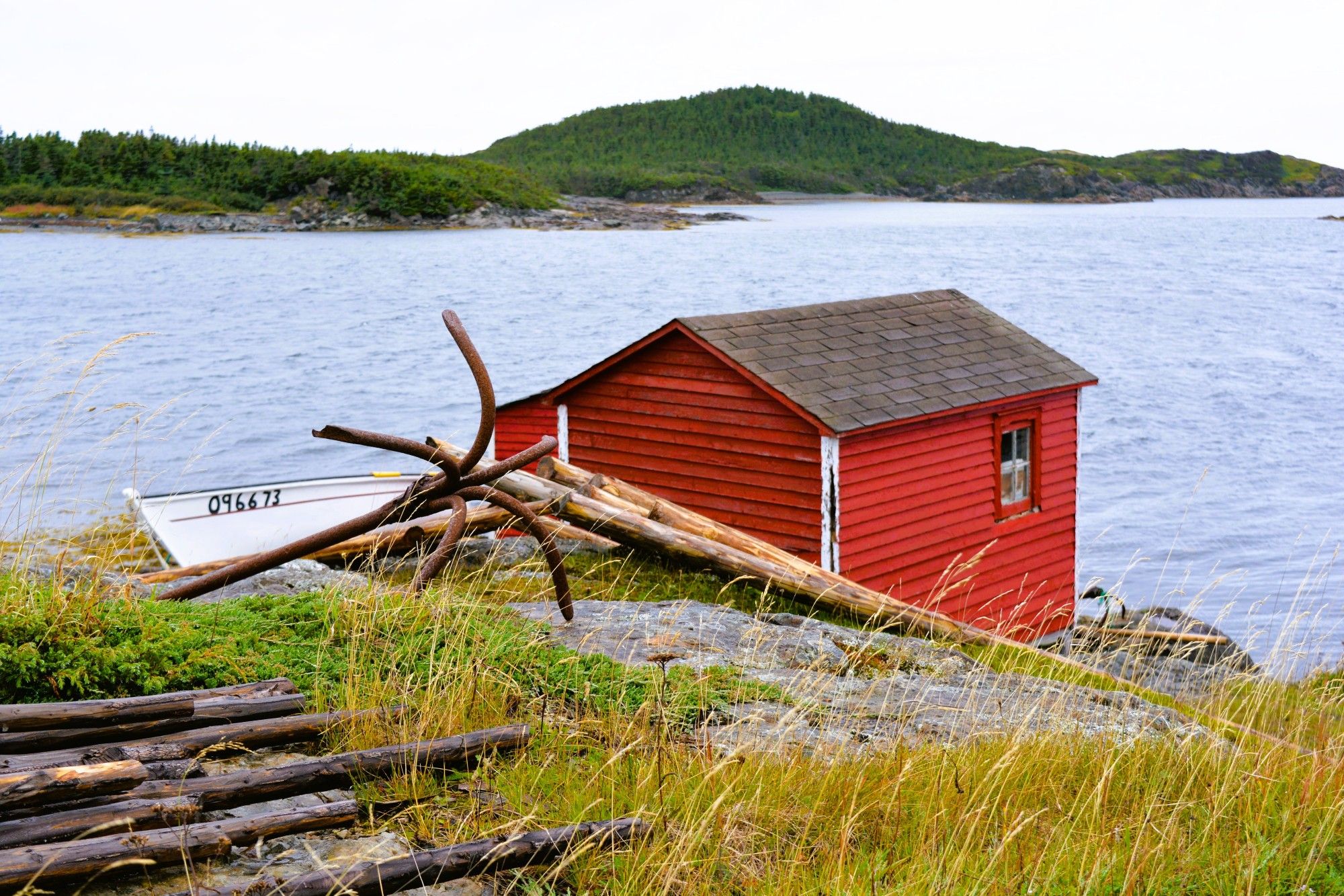 A red shed with a brown roof and a single window on its side, is striking against the grey of the ocean that is bordered by green hills on the far shore. To the side of the shed is a white boat nearing a set of numbers. There are also a number of logs and yellow/green reefs strewed around.