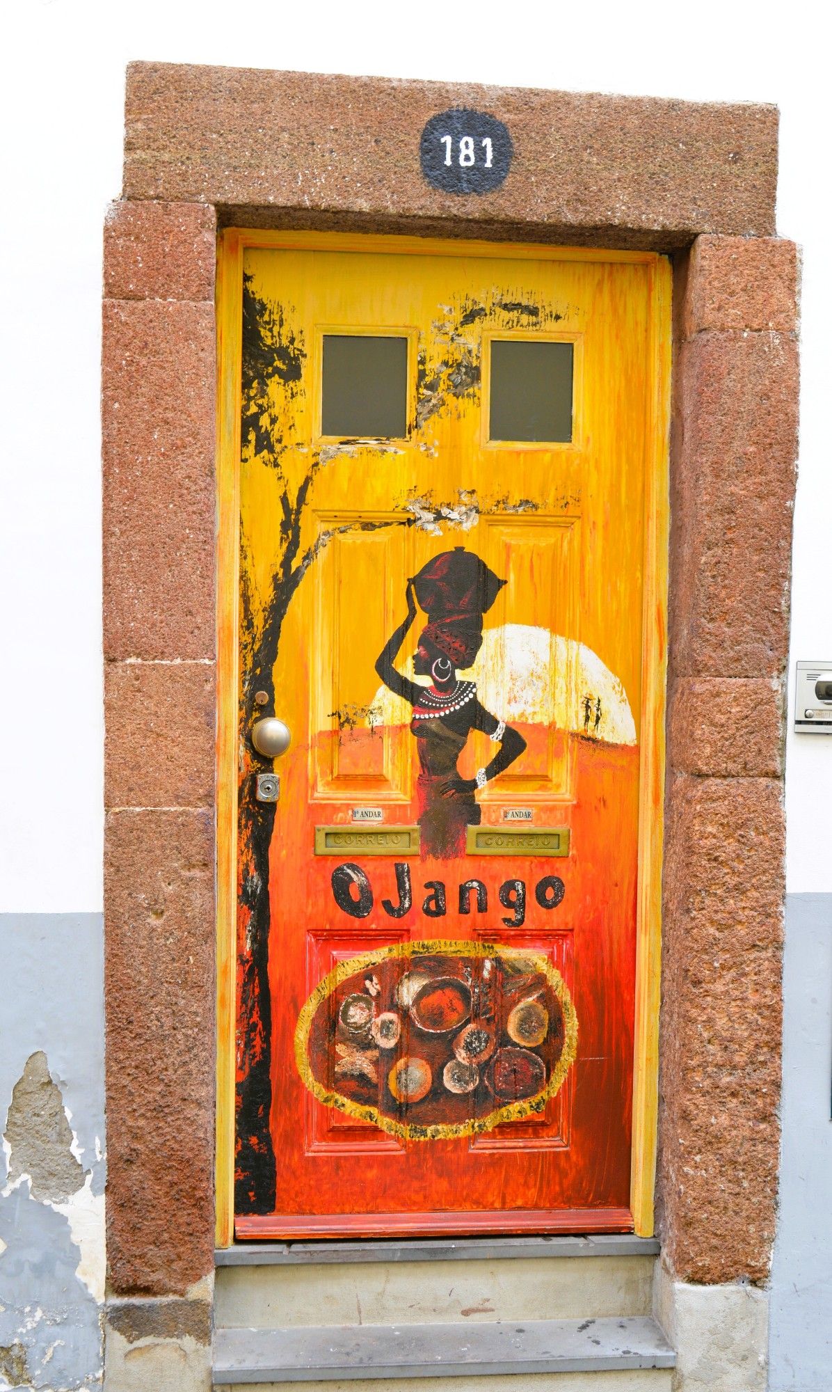 A painted door in yellow and orange, with the word Jango across it. In the middle a painting of an African woman poses proudly with a basket on her head. An assortment of dishes take over the lower half. The branch of a tree runs up the left hand side and branches out between two square windows at the top where the number 181 is displayed.