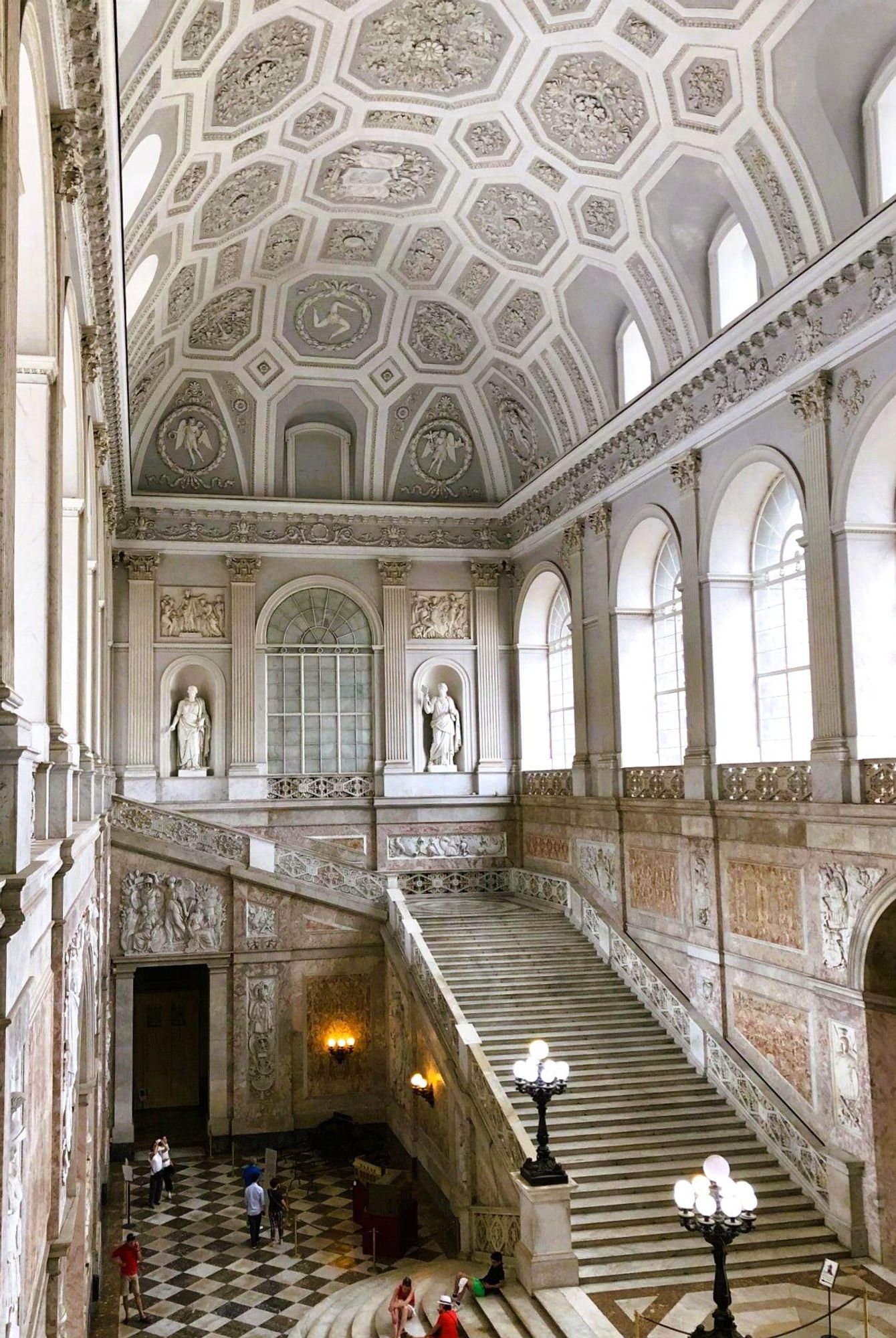 The other side of a sweeping marble staircase inside a palace. There are statues in alcoves in the walls, lamp posts with multiple globes, as well ornate decor on the walls and ceiling, and large arched windows letting in the light.