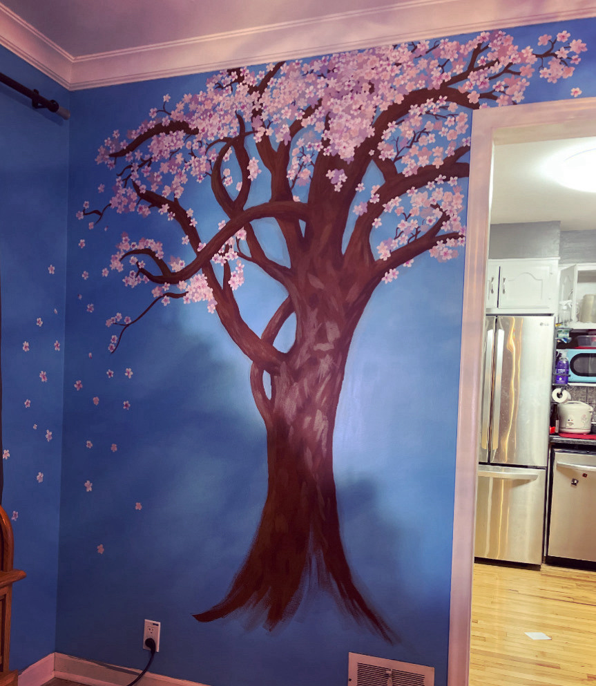 Painting on a sky blue wall with white trim of a twisting cherry blossom tree. The hundreds of flowers are in a multitude of pinks and light purple. Some are even cascading down to the ground on the left side as if blown by the wind.