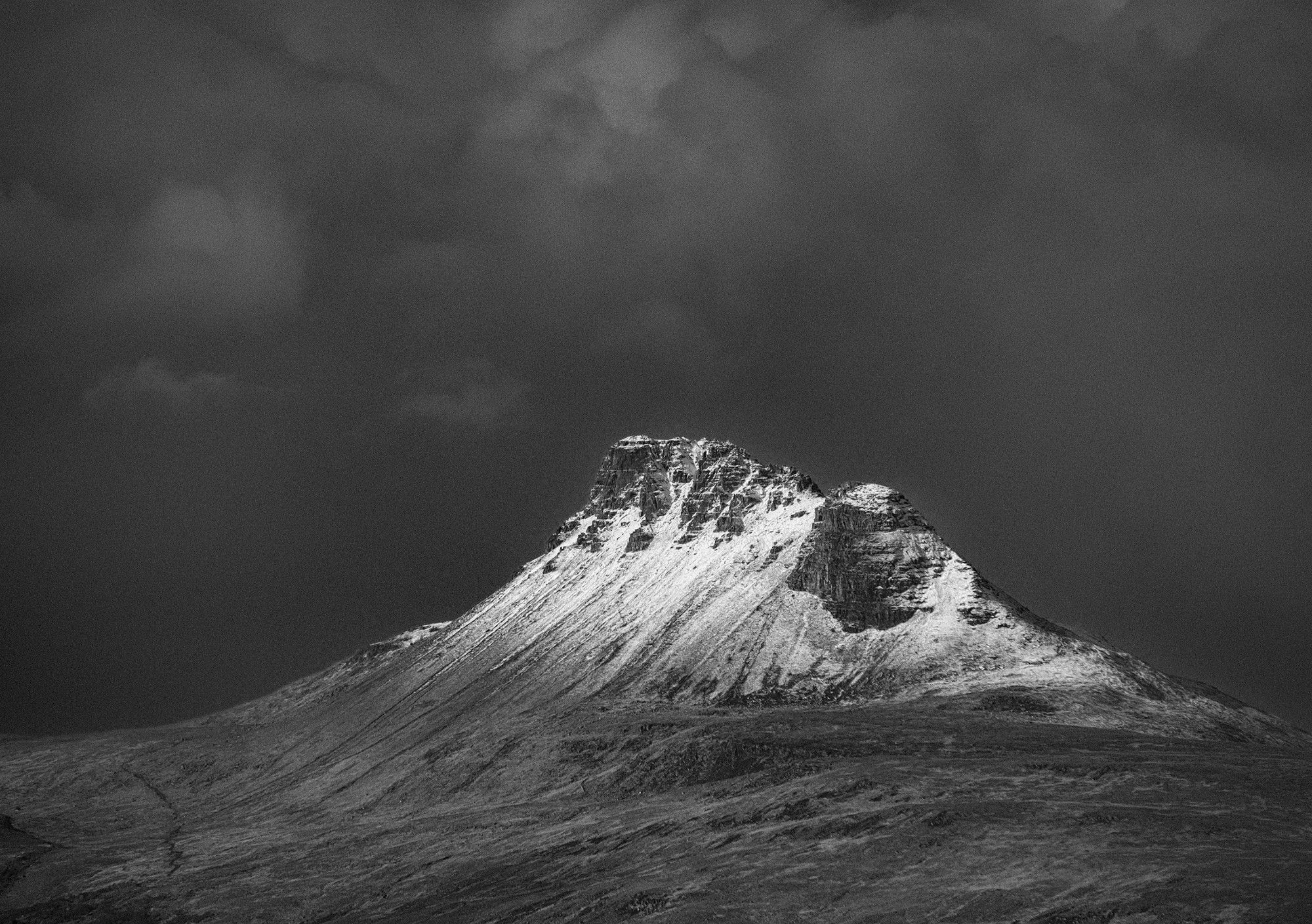 A black and white photo of a snow dusted Stac Pollaidh, the distinctive upper rocky layers giving way to the long sloping scree slopes. It stands high above the surrounding land, which is below the snow line. In the background, a dark, ominous, snow filled sky, with indistinct clouds within provides a good contrast to the snowy mountain. Highlands, Scotland.

Photo by and copyright of Paul Henni.