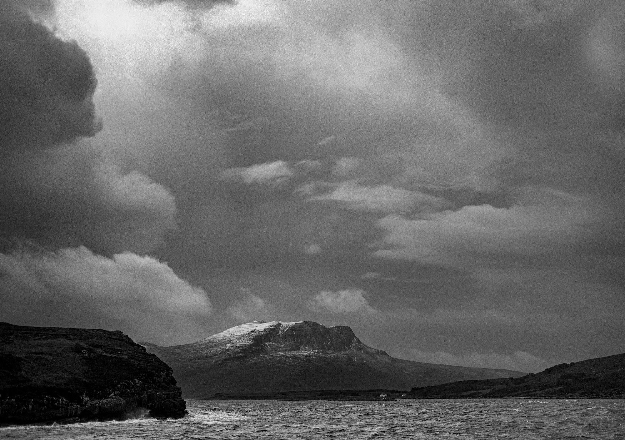 A black and white photo. Isle Martin and the sea are low in the foreground, above them the snow dusted Beinn Ghobhlach appears in a gap in the dark clouds at the entrance to Loch Broom, near Ullapool, Wester Ross, Scottish Highlands.

Photo by and copyright of Lynn Henni.