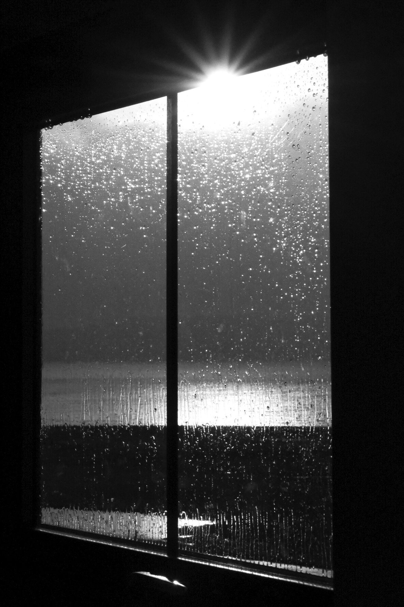 A black and white photo looking out of a wet window to Upper Loch Torridon, which is partially in bright sunshine, with looming clouds and shafts of light above it. Torridon, Wester Ross, Scottish Highlands.

Photo by and copyright of Paul Henni.