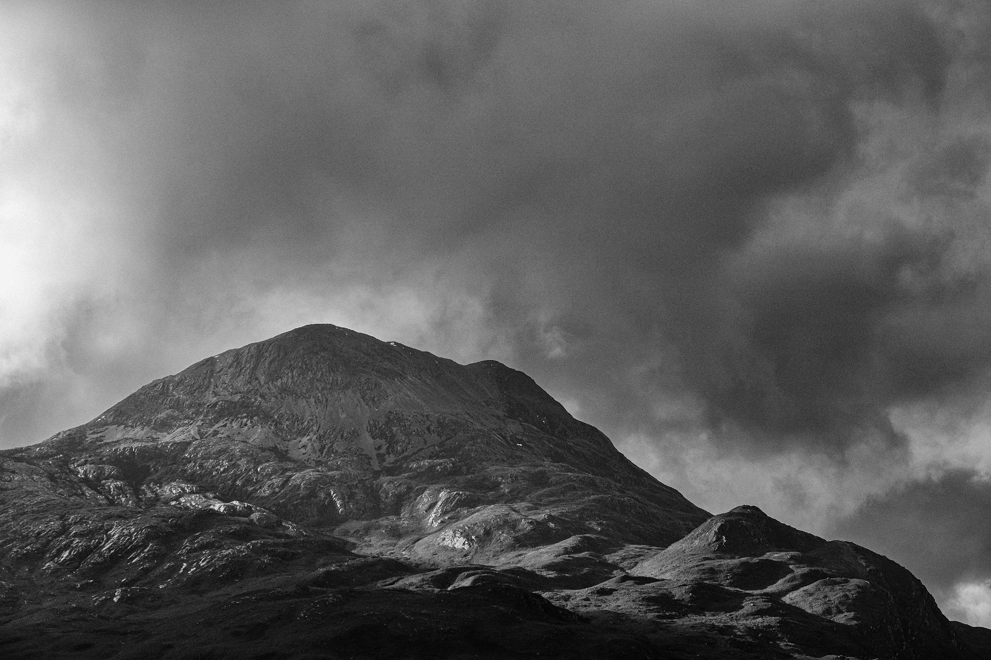 A black and white photo of Sgùrr Dubh or The Black Peak, with Craig Dubh or Black Rock below, as they as it rise above Loch Coulin in the Torridon area. Mixed clouds are slowly being illuminated by the sun as it cuts across the scene from the left. The peak and bottom of the frame are dark, whilst a shaft of sunlight brightens the hummocky lower slopes of the mountain.  Wester Ross, Highlands And Islands, Scotland.

Photo by and copyright of Paul Henni.