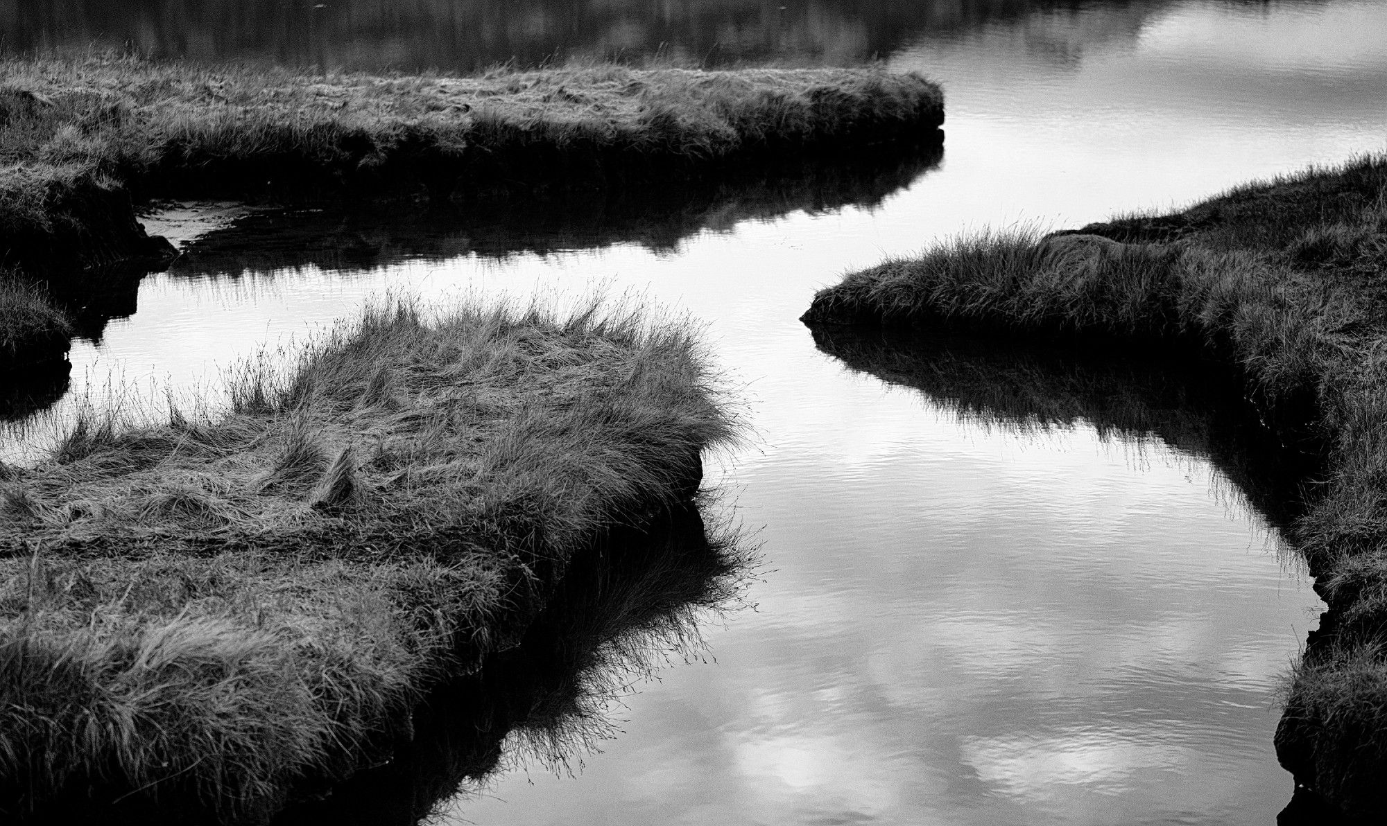 A black and white photo of mirror the like waters of the salt marsh on the shores of Upper Loch Torridon, just in front of Annat. Fingers of grassy land reach in from the edges of the frame. Annat, Torridon, Wester Ross, Highlands, Scotland.

Photo by and copyright of Paul Henni.