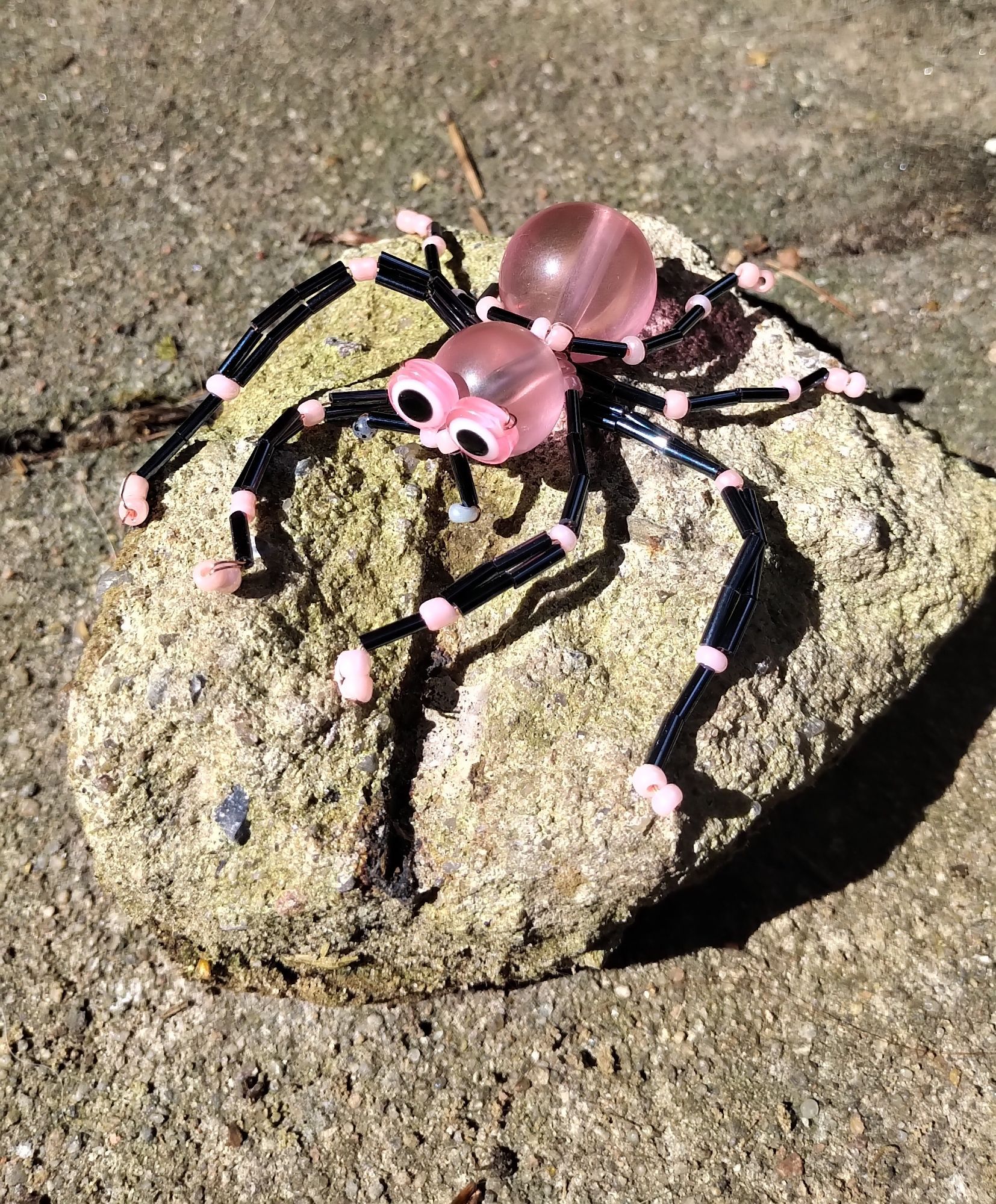 Eine Spinne aus Perlen gebastelt. Der Körper und die Augen sind rosa. Die Beine schwarz mit rosafarbenen Gelenken und Füßen. Sie sitzt auf einem etwa faustgroßen Stein.