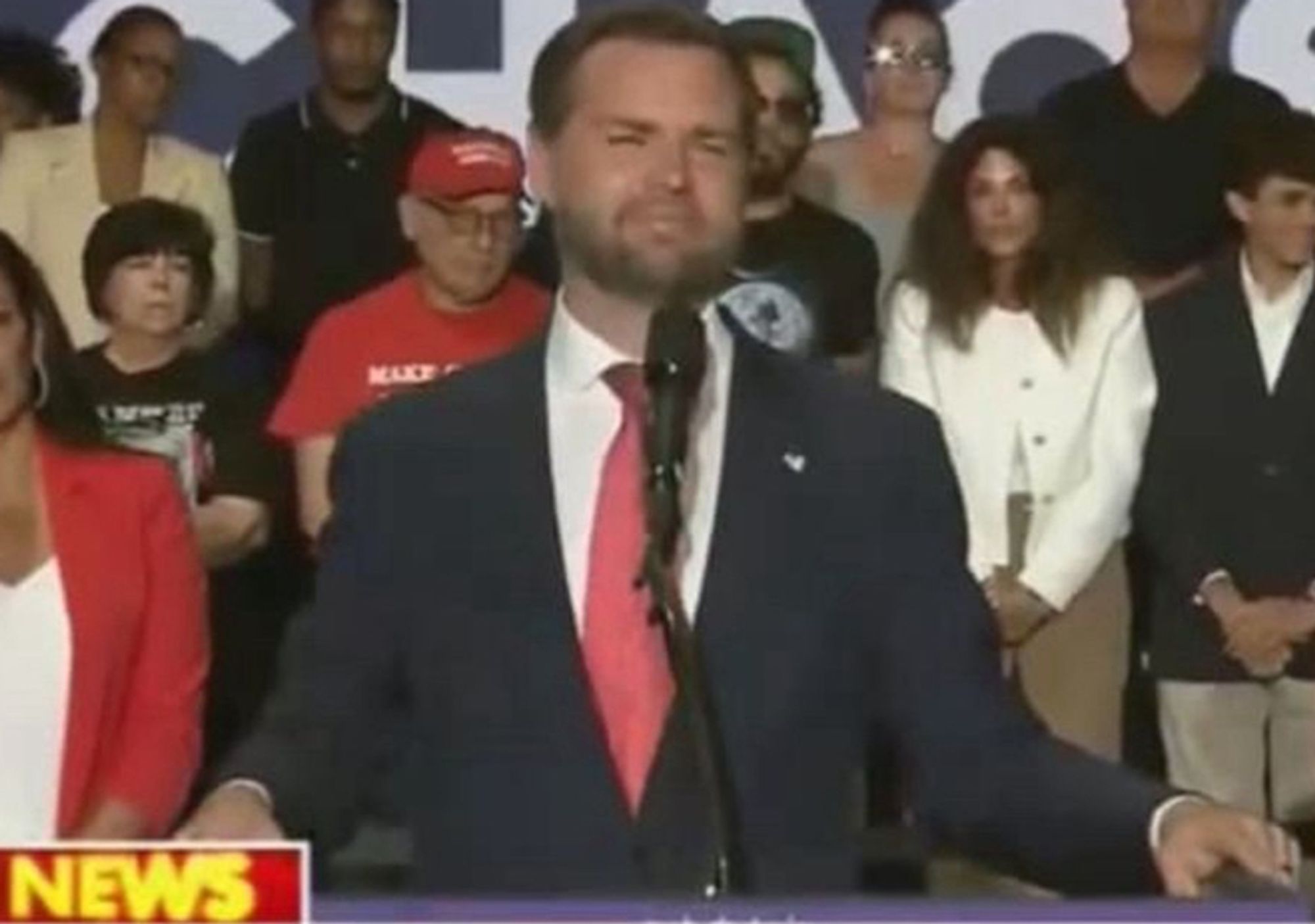 A snip from the above picture of JD Vance smirking at the camera standing in front of the microphone. The word NEWS from the Fox news "BREAKING NEWS" chyron is in the bottom right corner