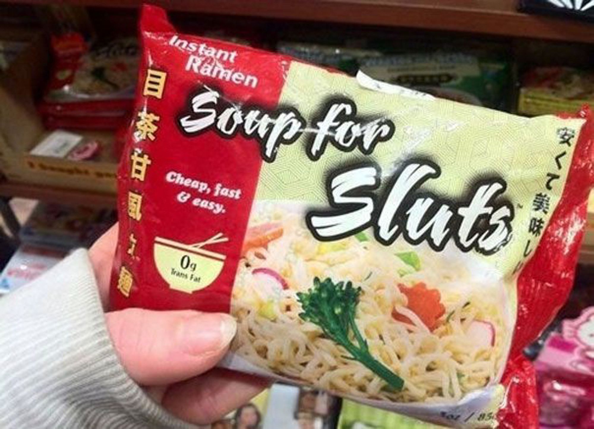 A woman's hand holding up a red-bordered pack of instant ramen called "Soup for Sluts". It says 'Cheap, fast & easy.' below that. The picture of the ramen on the package has fancy radishes and carrots in it and looks very tasty. 

The package also informs you it has 0g trans fat!