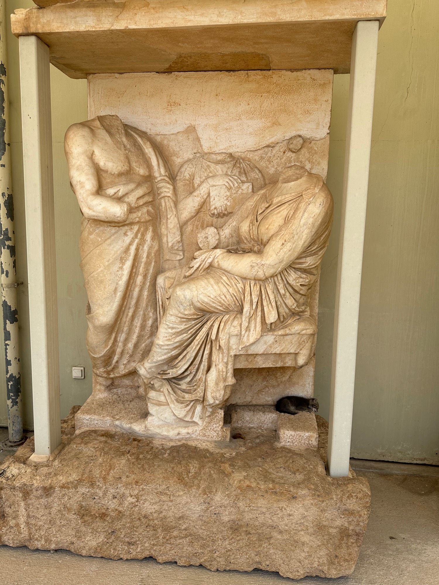 Marble funerary stele containing a steamed man, and a standing man both in high relief, with a standing woman in the background in low relief, on a small ledge below the seated man is a sleeping kitten (real)