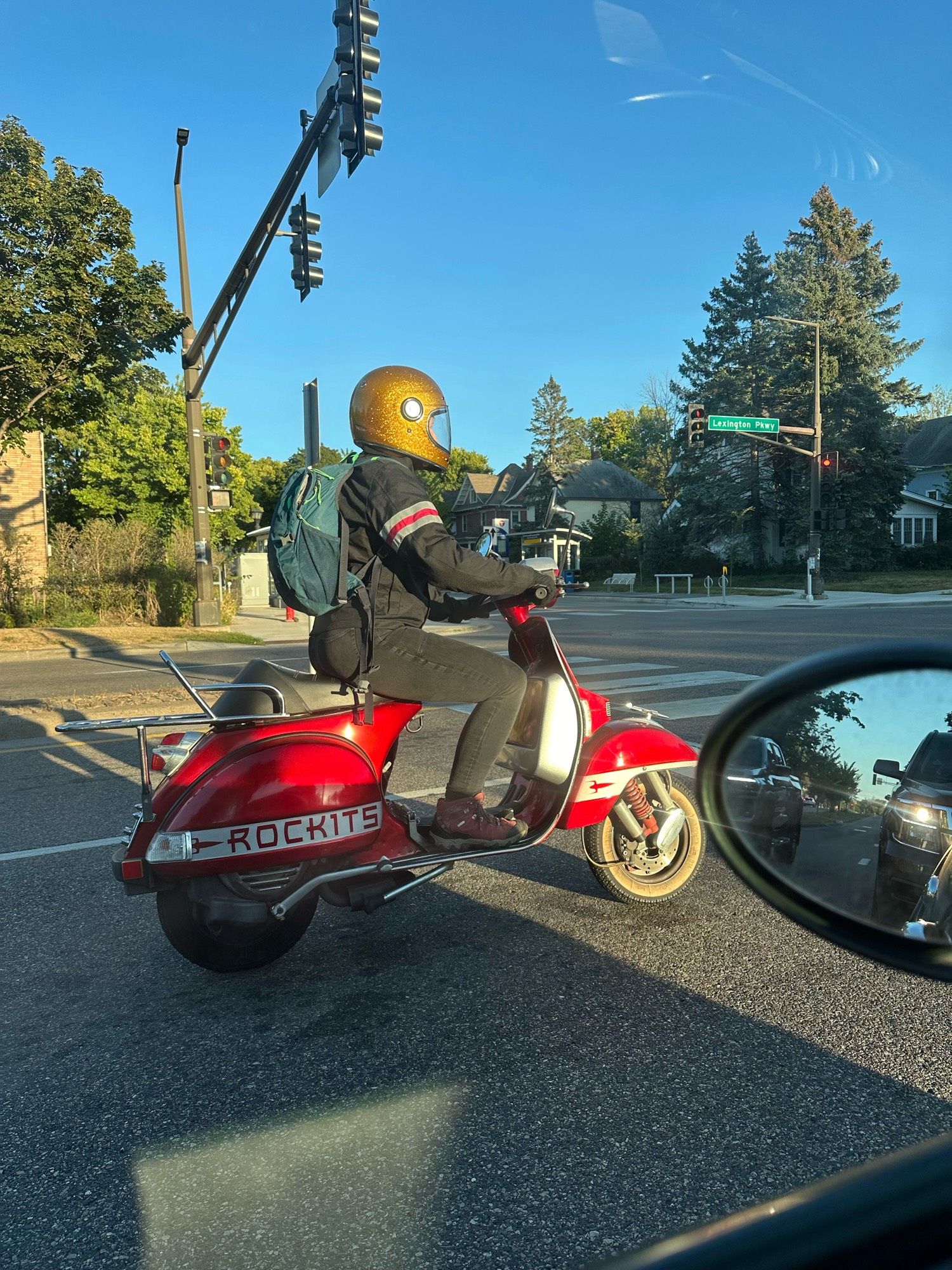 A person riding a red scooter with a silver stripe along the wheel well with the word “Rockits”, a roller derby team name. The rider is wearing a glittery gold full face helmet, a teal backpack, black pants, raspberry colored hiking boots, and a black riding jacket with a silver and raspberry stripe on the arm.