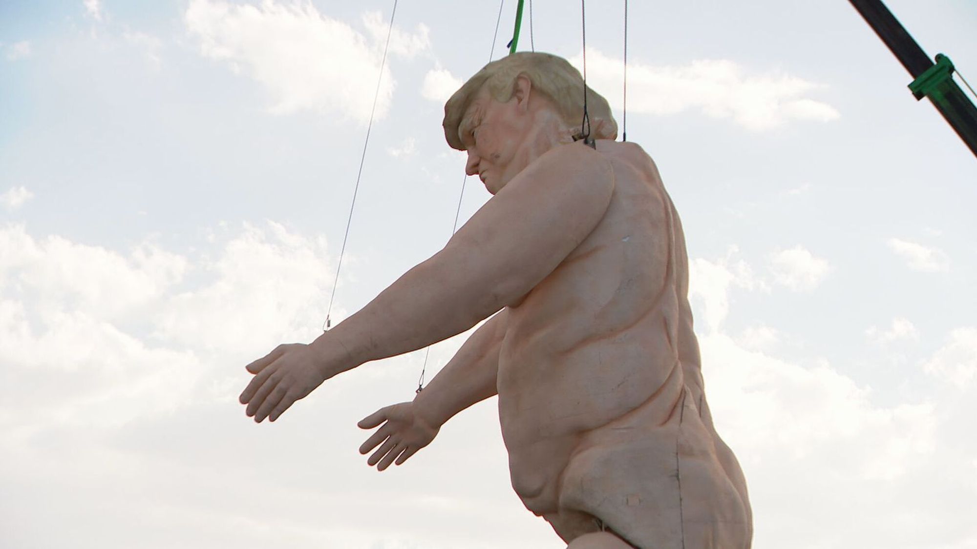 A large statue of a nude Donald Trump being hoisted by a crane