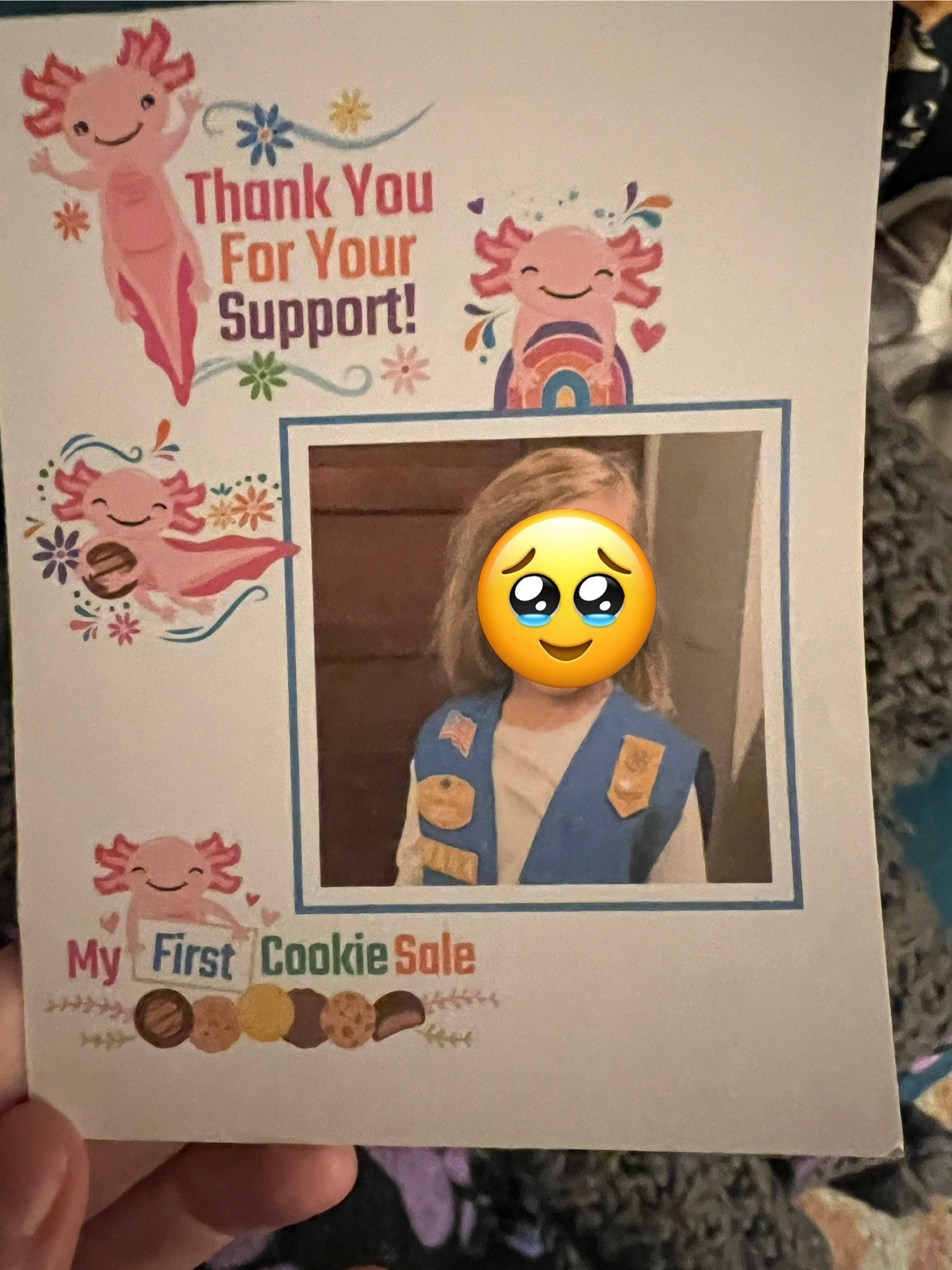 The front of a thank you card. There is a picture of the scout on the right & the rest is decorated with pink axolotls that say “Thank you for your support” and “My first cookie sale”. I’ve put a smilie face over the child’s face for privacy reasons.  