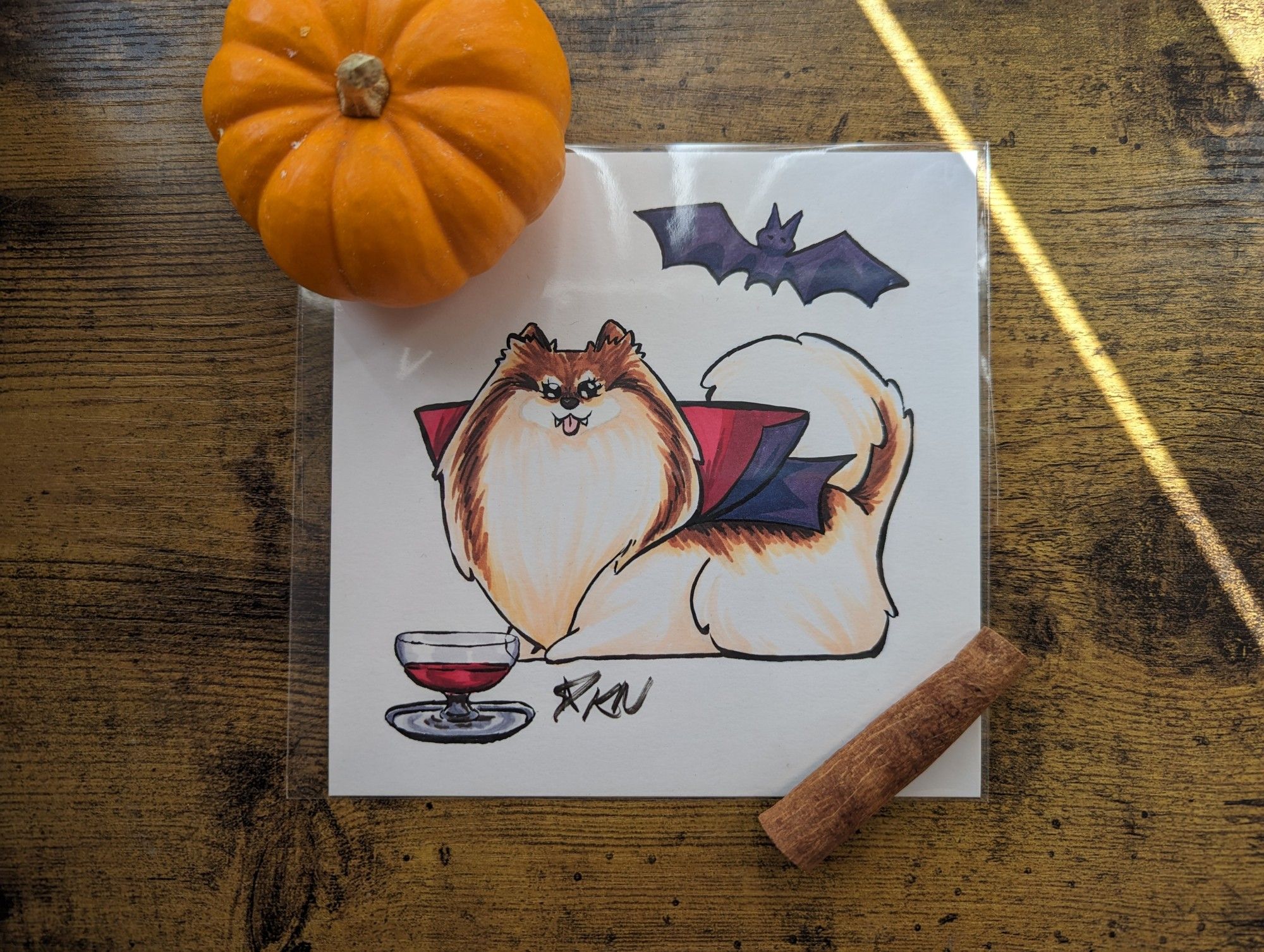 An illustration of a white and tan pomeranian sitting prim and proper wearing a vampire cape. She has two little vampire fangs poking out around her toungue and a chalice of juice in front of her, there's a bat flying overhead.