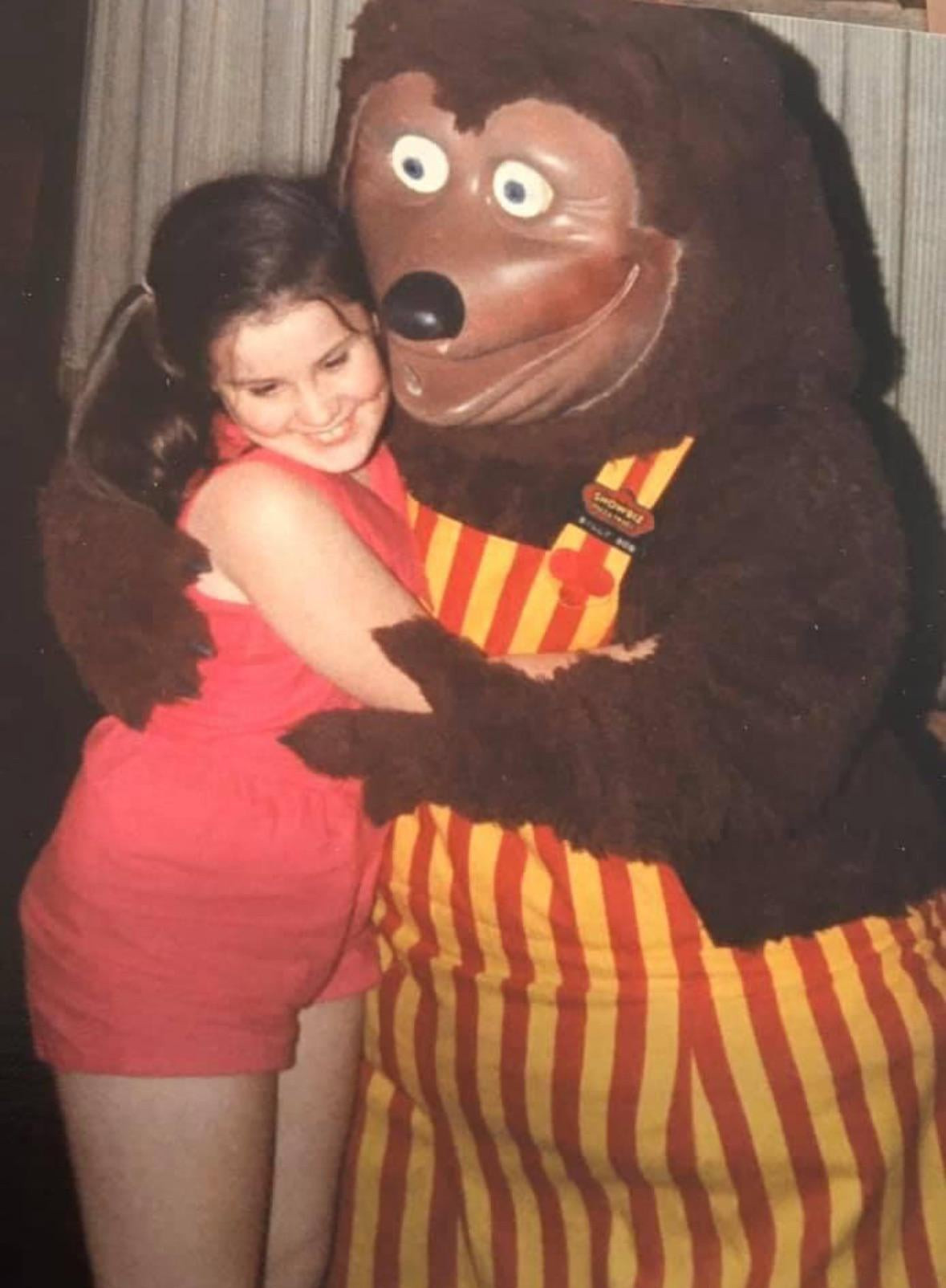 A person hugging a cursed bear costume that's wearing red and yellow striped overalls